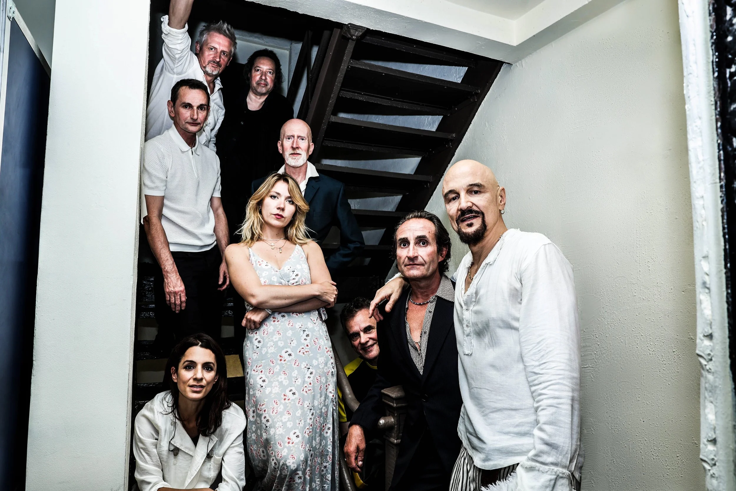 A group of nine diverse adults, including women and men of various ages, gathered on a staircase in an indoor setting. They are looking at the camera with neutral to serious expressions. The background features plain walls and part of a staircase.