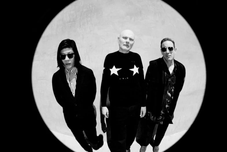 Three people standing in a line with a plain wall background, viewed through a circular fisheye lens, wearing dark clothing and sunglasses, black and white photo.