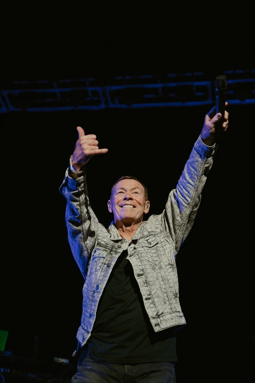 A man smiling and holding a microphone in one hand while raising the other hand in a 'call me' gesture on a dark stage.