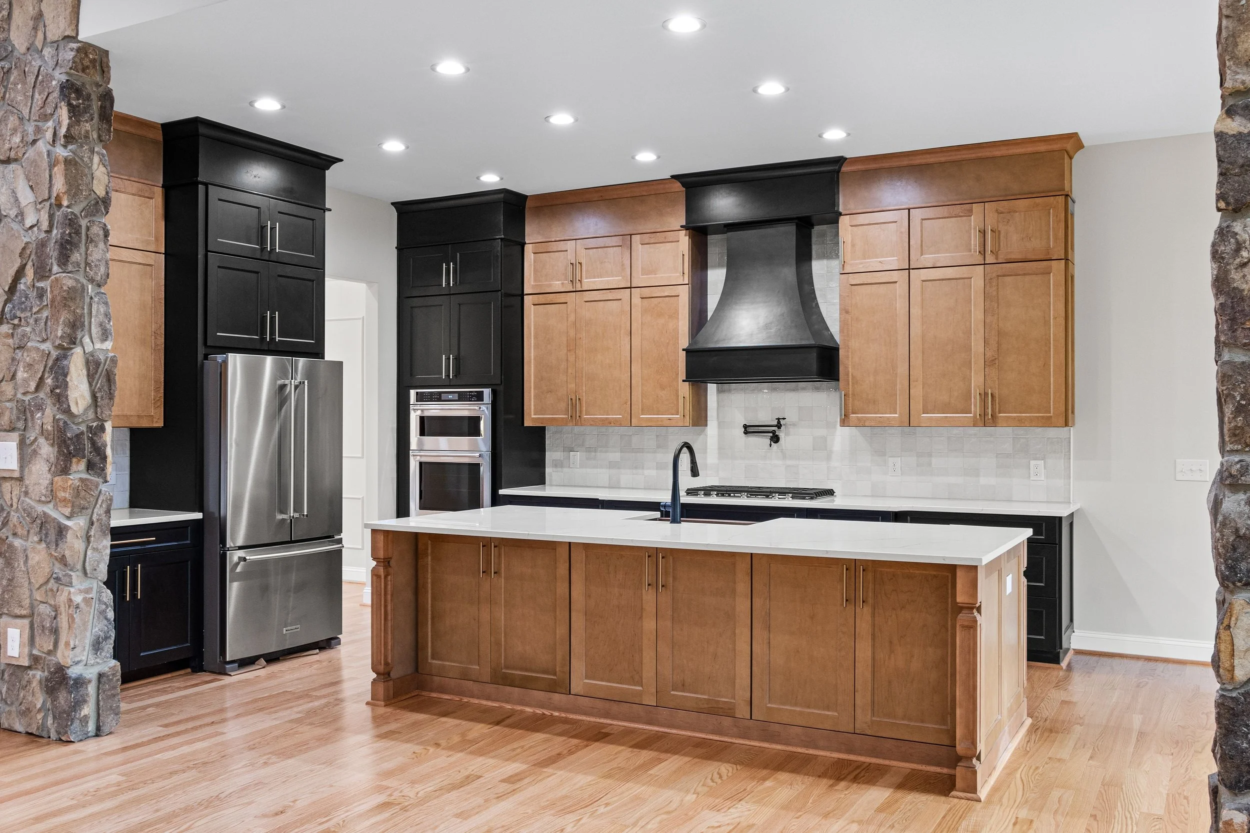 Modern kitchen with black and natural wood cabinets, stainless steel refrigerator, black range hood, white countertops, and a stone wall on both sides.