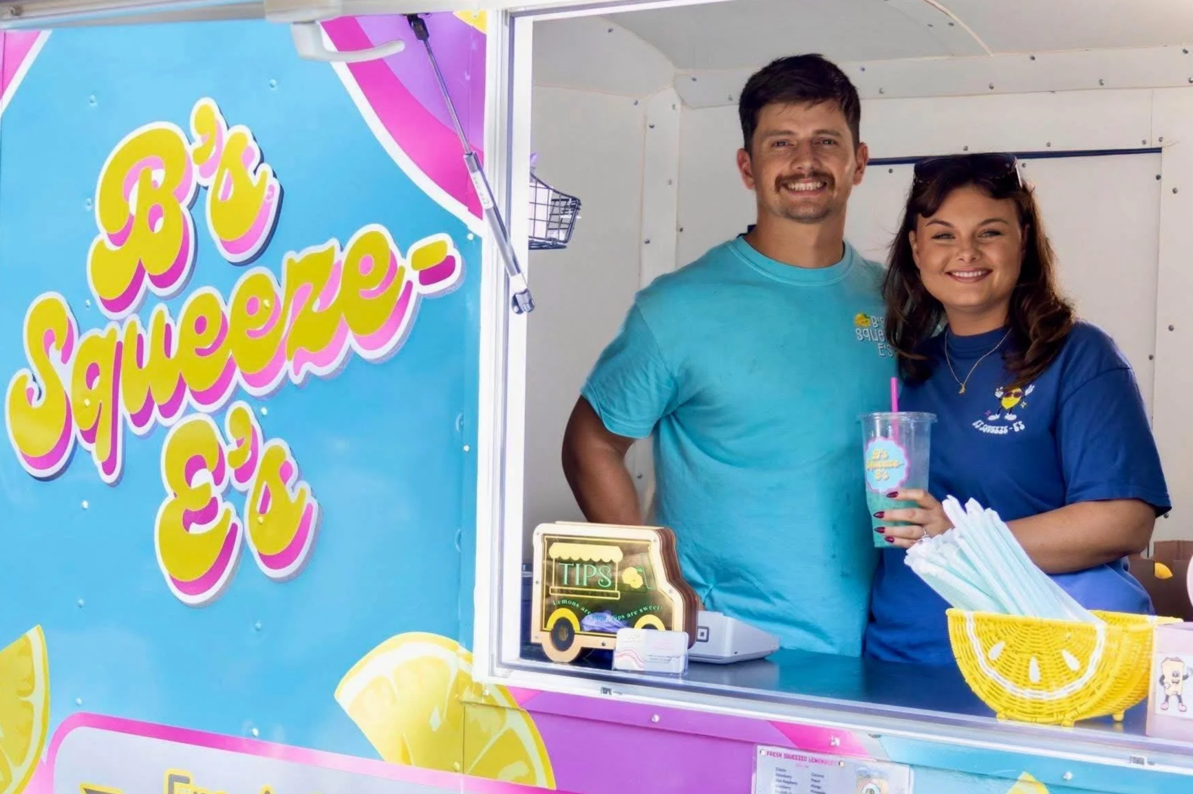 A man and woman smiling at a lemonade stand with a colorful sign that reads 'B's Squeeze-E's and a container of lemonade with a lemon graphic. The woman is holding a clear cup with a pink straw, and there are yellow and white items on the counter.