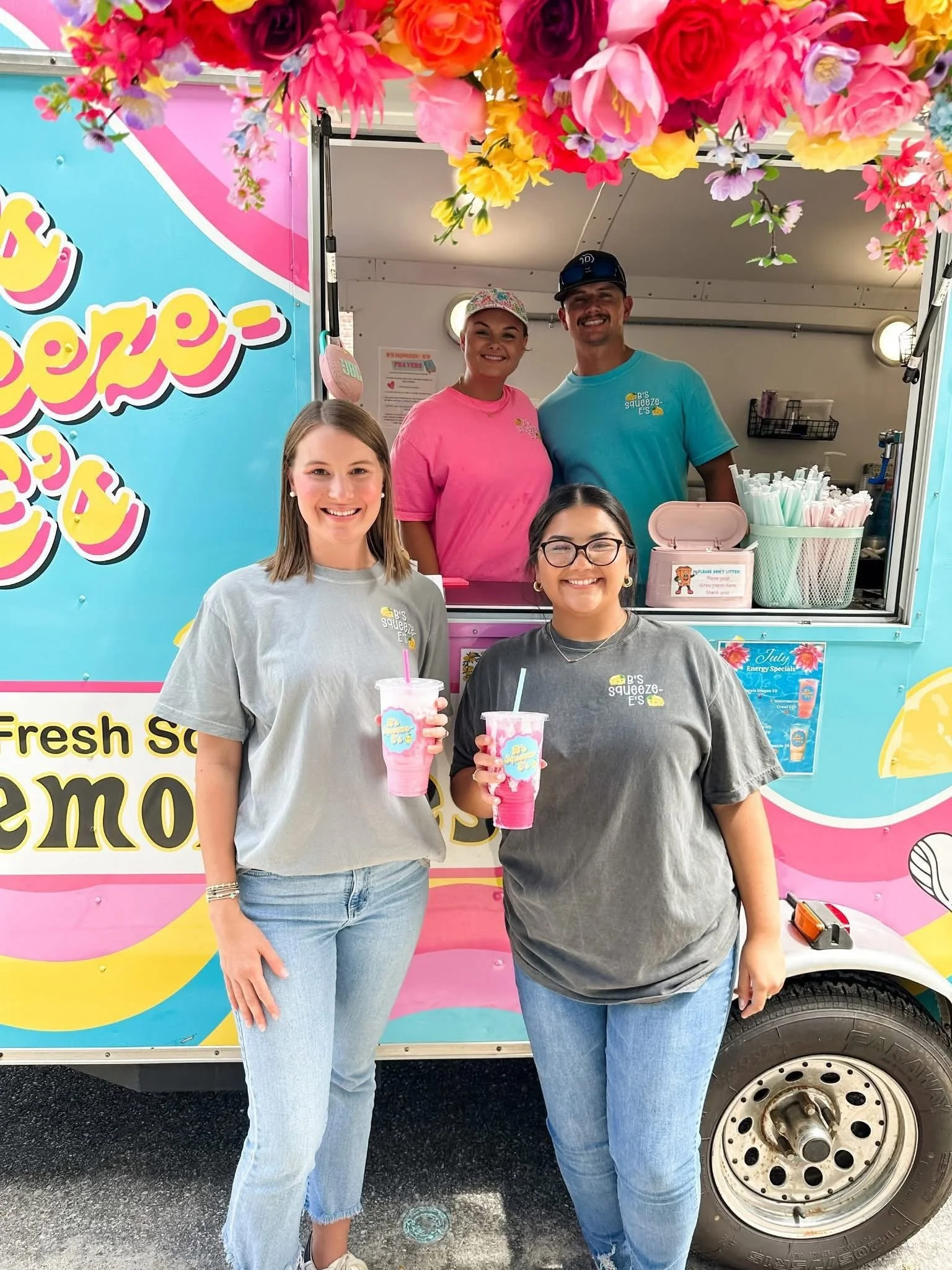 Four women standing in front of a colorful ice cream truck decorated with pink and yellow flowers, holding pink and white ice cream drinks, smiling at the camera.