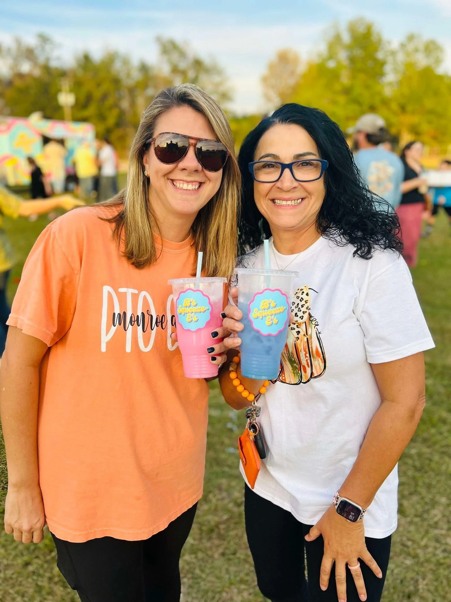 Two smiling women holding drinks at an outdoor event, with food trucks and trees in the background.