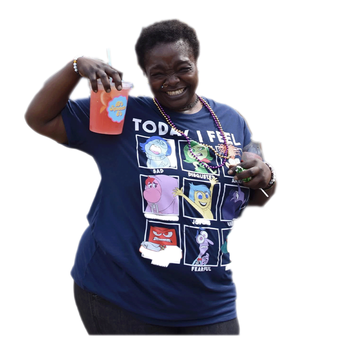 A woman with dark skin, short hair, and a nose piercing smiling joyfully shows off her colorful drink. She wears a navy blue T-shirt featuring various cartoon characters expressing different emotions, and necklaces with beads. The background is black.