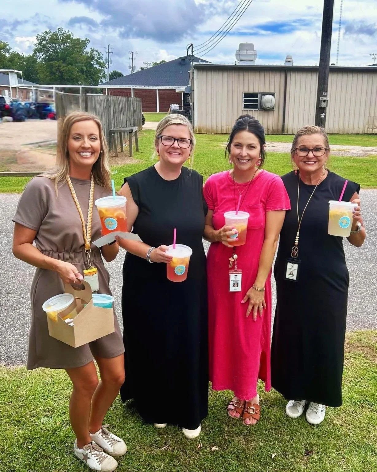 Four women standing on grass outdoors, smiling, holding drinks, and facing the camera. Background shows a small building, parked cars, utility poles, and a partly cloudy sky.