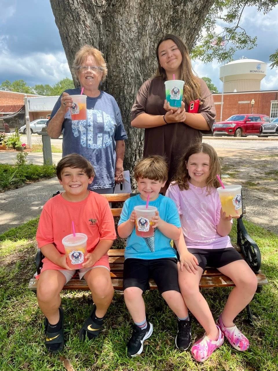 Five children and two women outdoors in front of a large tree, holding colorful drinks with straws. The children are sitting on a wooden bench, smiling, with three wearing pink, blue, and peach shirts. The women are standing behind, smiling, one wearing glasses and a blue shirt, the other in a brown shirt. The background features a parking lot with cars and a water tower.