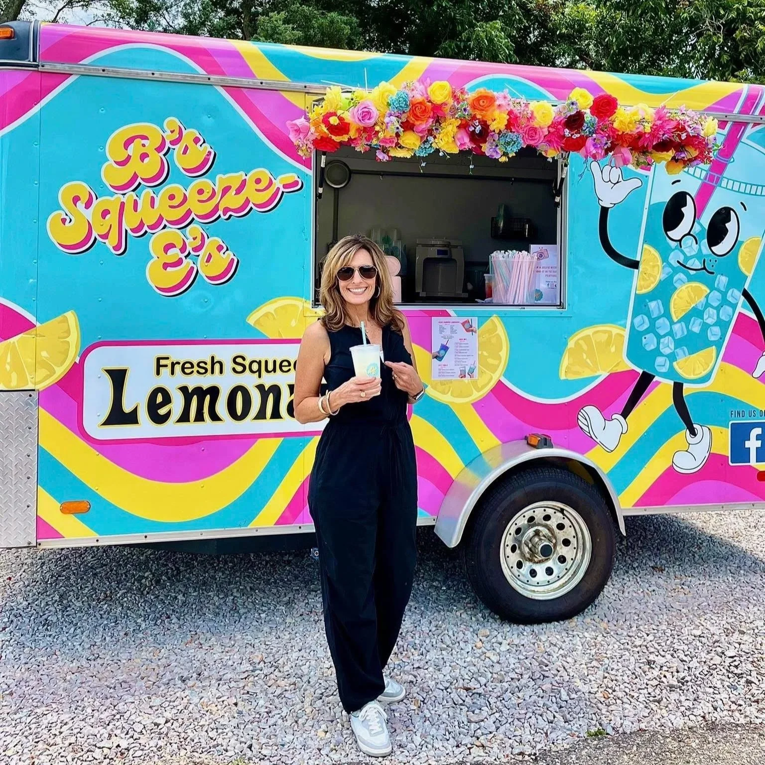 A woman with blonde hair wearing sunglasses and a black sleeveless jumpsuit, smiling and holding a lemon-themed drink, standing in front of a colorful lemon-flavored beverage truck decorated with flowers.