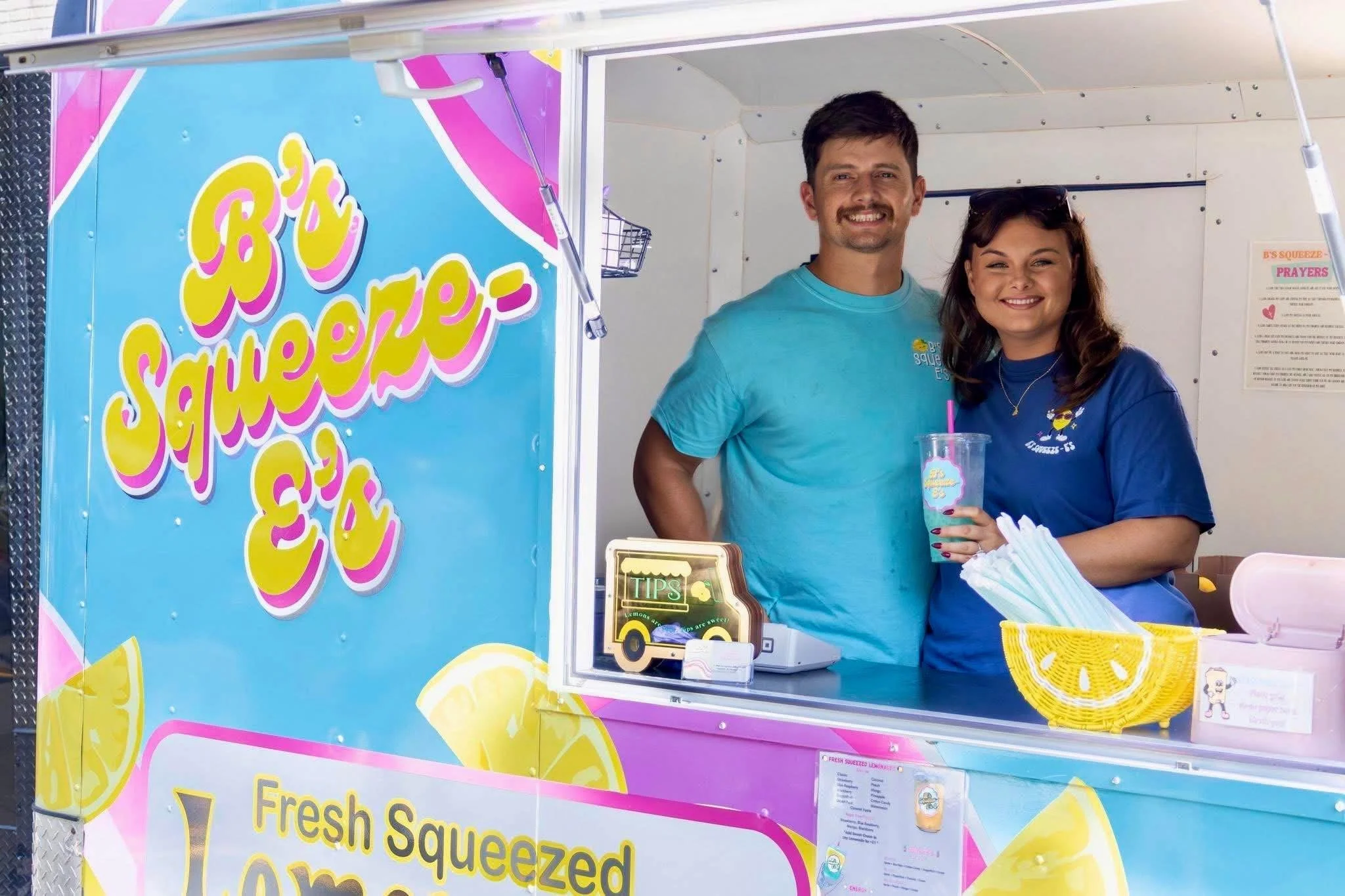 A photo of a smiling man and woman standing inside a bright pink and blue lemon-shaped lemonade stand booth. The booth has a sign that reads "B's Squeeze" and offers fresh squeezed lemonade. The woman is holding a clear plastic cup with a pink straw, and there is a basket of yellow straws on the counter. The man is wearing a teal t-shirt, and the woman is wearing a matching blue t-shirt with the stand's logo.