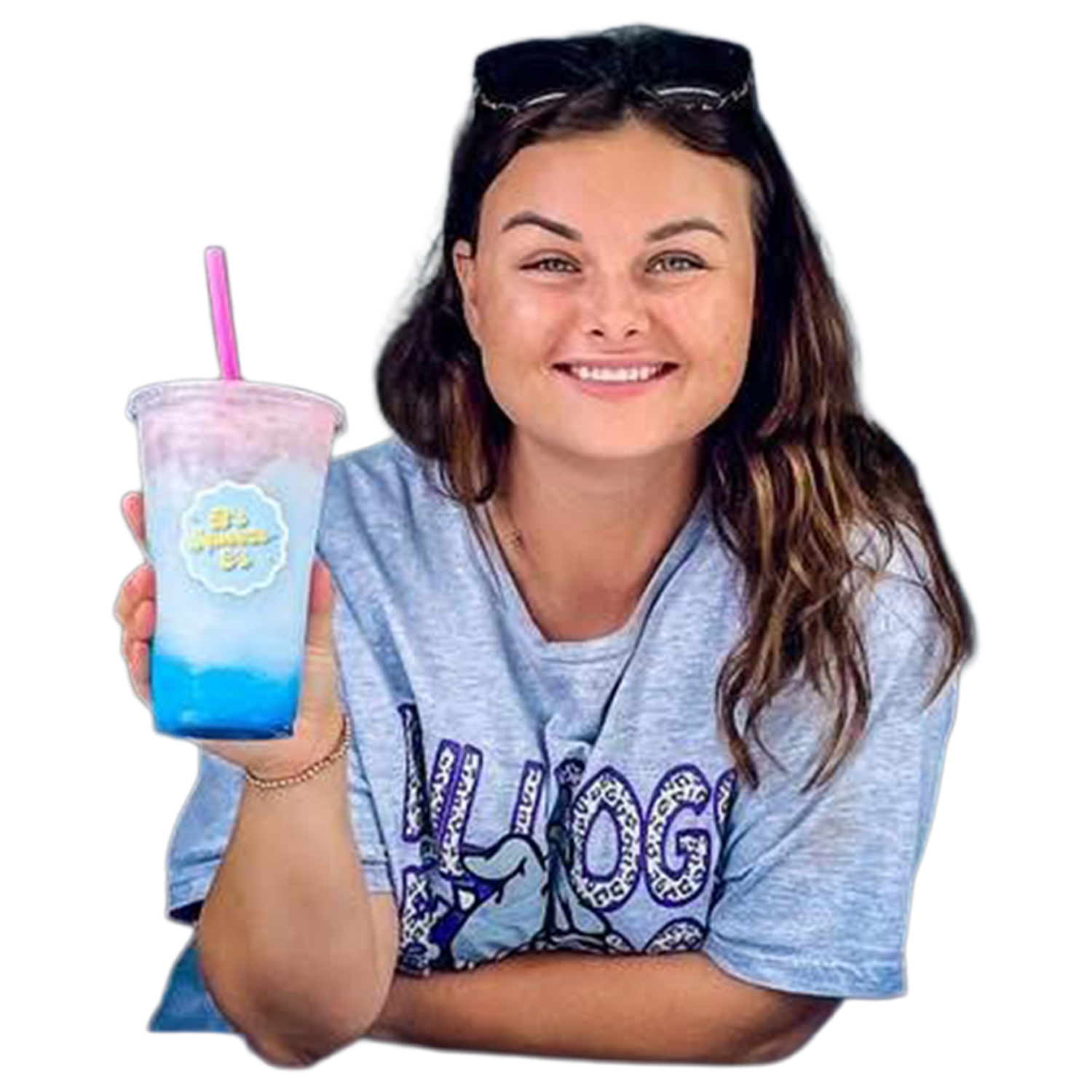 A young woman with long brown hair, wearing sunglasses on her head and a gray T-shirt with purple text, is smiling and holding a colorful blue and pink drink in a clear plastic cup with a pink straw.