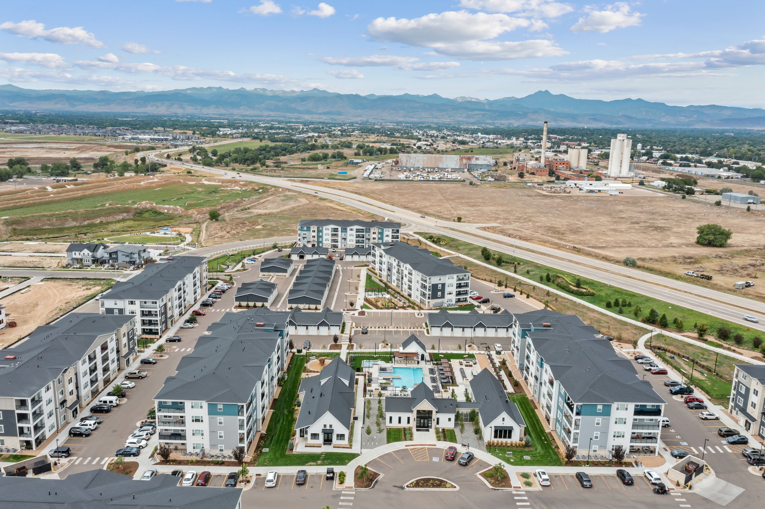 Aerial view of a residential area with modern townhouses and apartment buildings, a swimming pool, parking lots, and a landscape with open fields, industrial buildings, and mountains in the background under a partly cloudy sky.