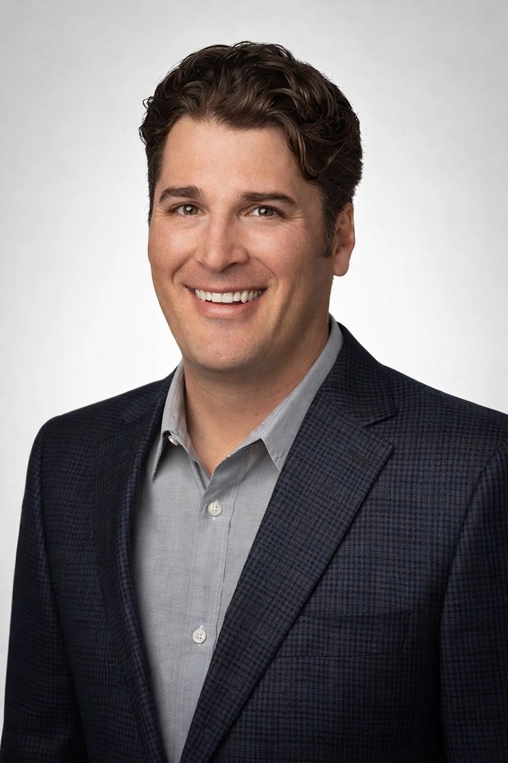 Professional headshot of a smiling man with dark wavy hair, wearing a checkered navy blazer and a light gray button-up shirt, against a plain white background.