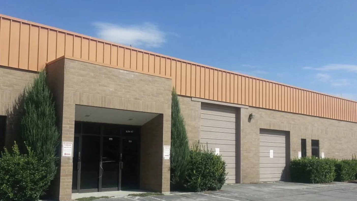 Exterior view of a large commercial or industrial building with brick walls, metal garage doors, and small windows, surrounded by shrubs and trees under a blue sky.