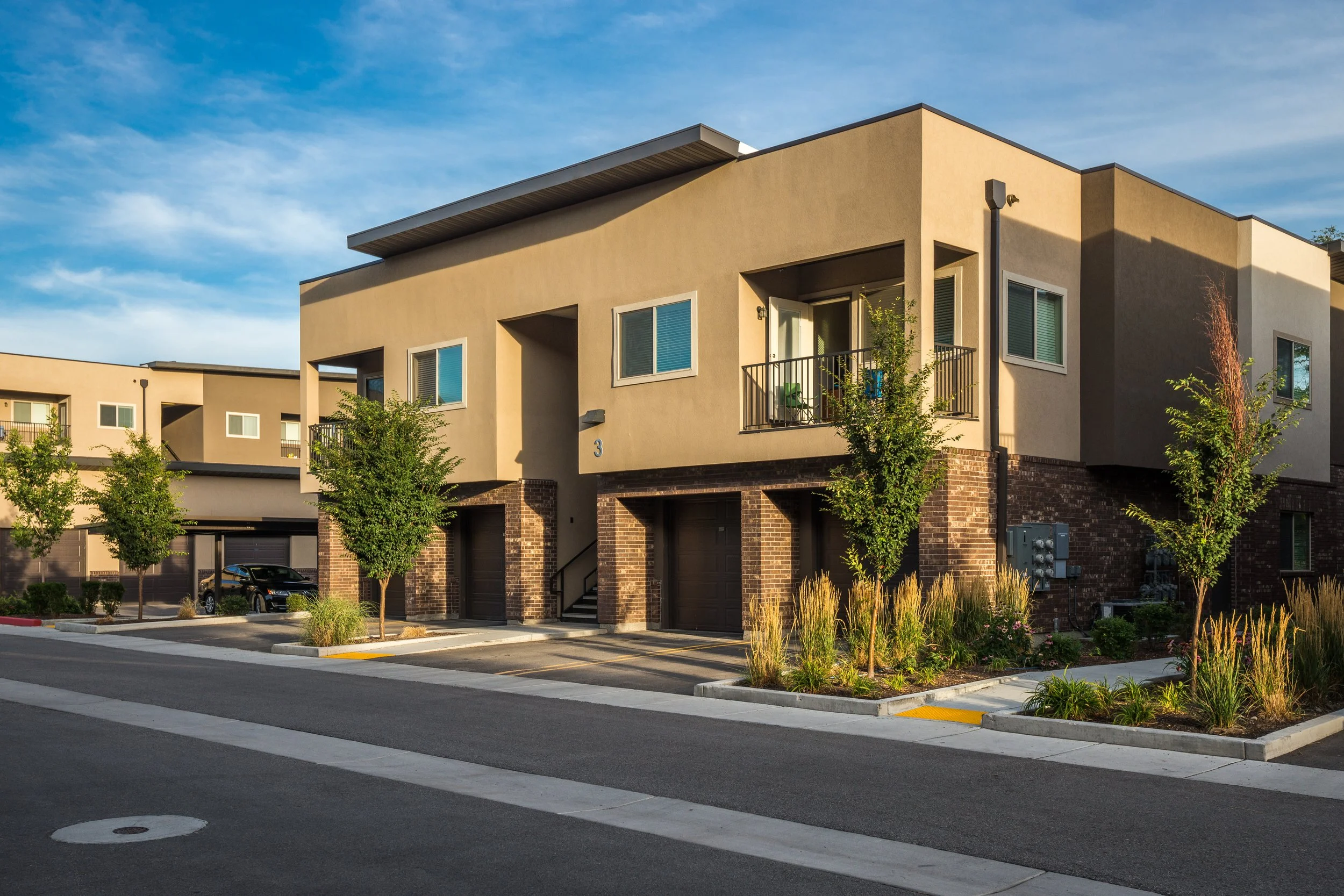 Residential apartment building with a tan stucco exterior, multiple windows, a balcony, and a garage on the ground floor. There are trees and a small landscaped area in front, under a partly cloudy sky.