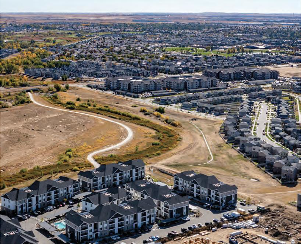 Aerial view of a residential area with apartment buildings, parking lots, and winding walking paths, surrounded by open land and more houses in the distance.