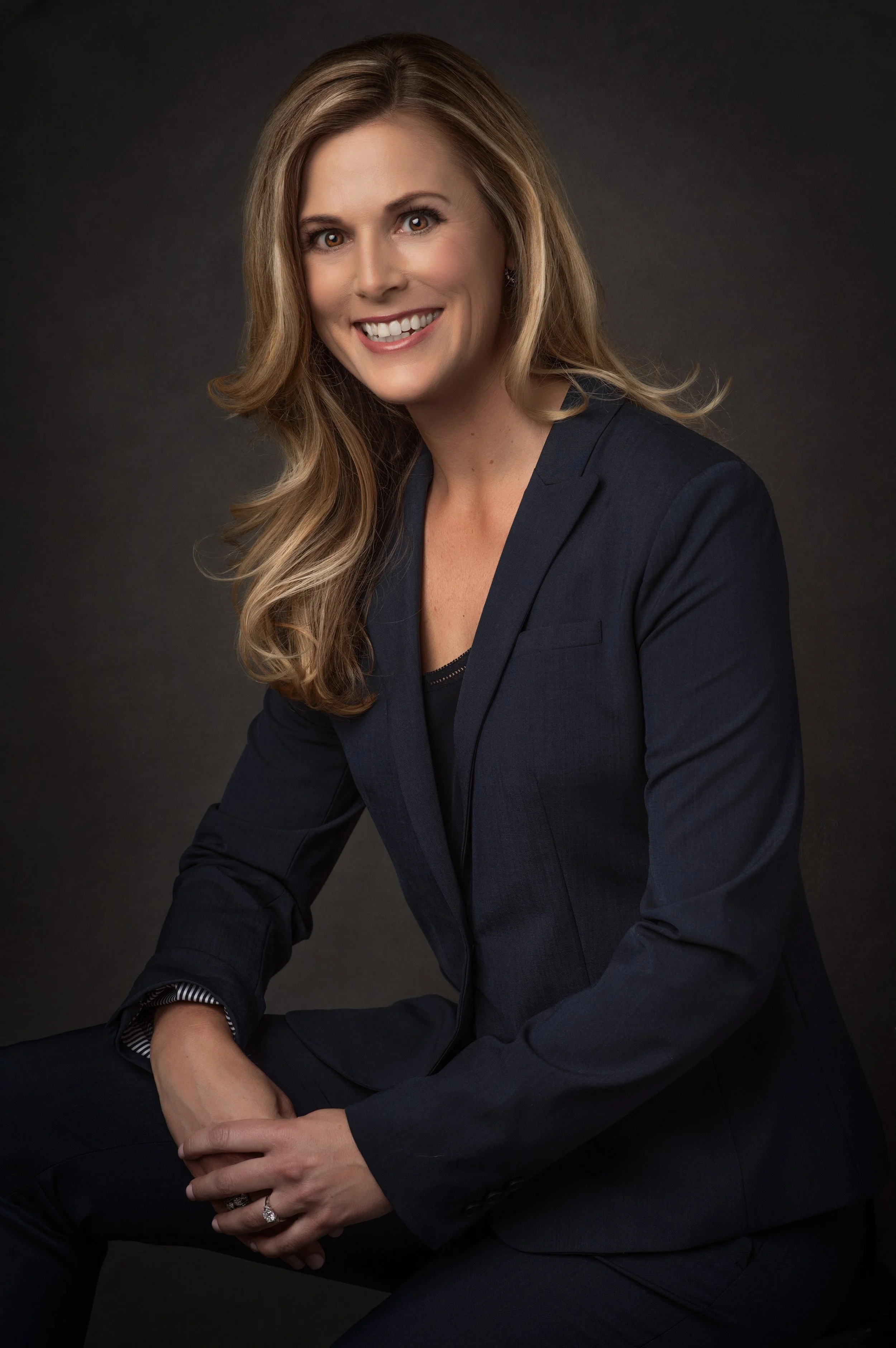 A woman with long, wavy blonde hair, wearing a dark navy blazer, smiling, sitting in front of a dark background.