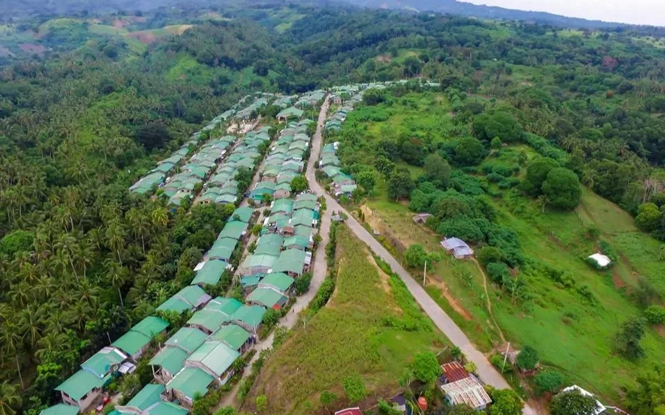 Aerial view of a rural village with numerous houses with green roofs surrounded by lush greenery and hills.
