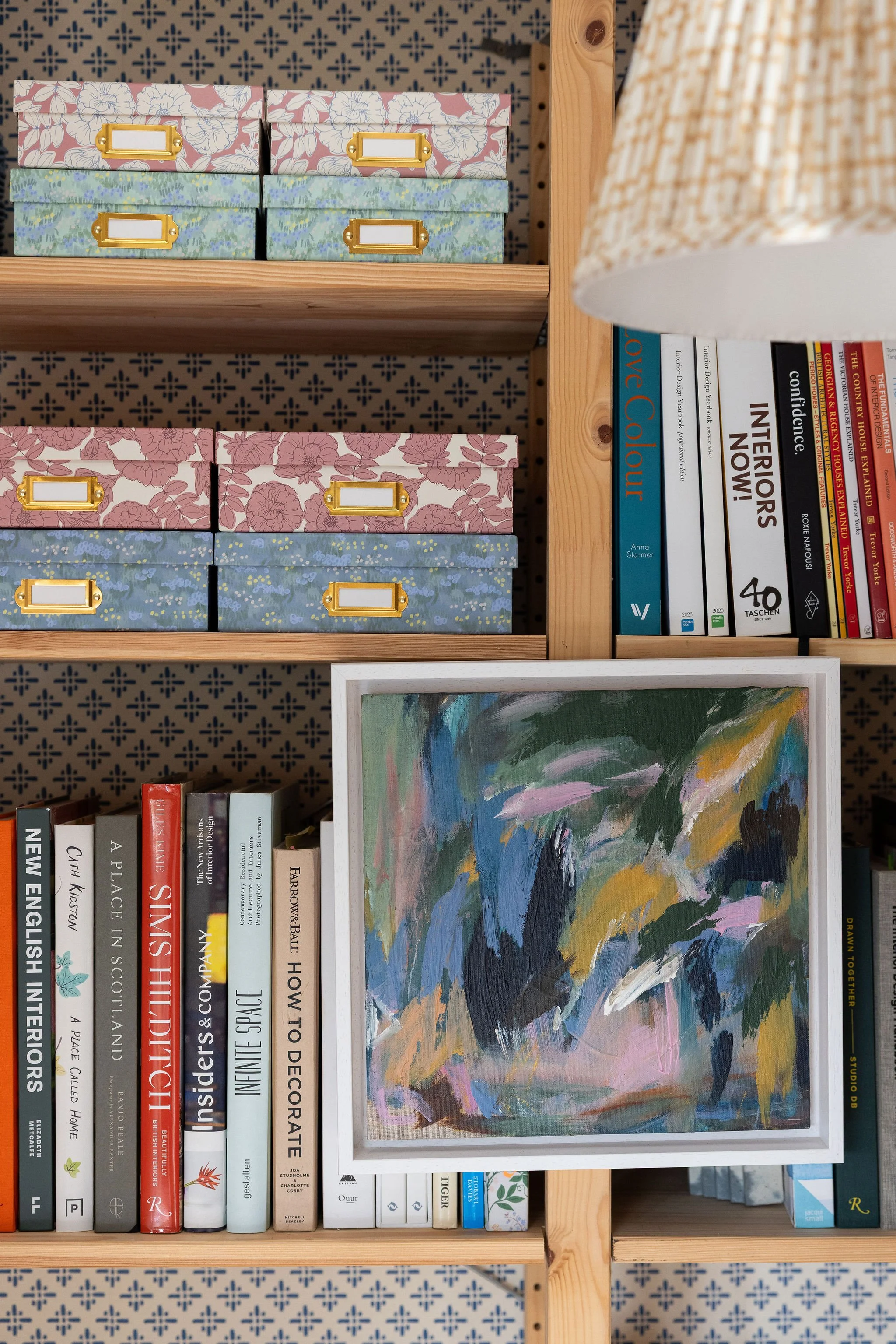 Bookshelf with two rows of books and decorative storage boxes on top. The shelf contains art books, interior design, and architecture titles, with some books titled 'The Interior Color,' 'Interiors Now!!,' and 'Infinite Space.' There is a framed abst