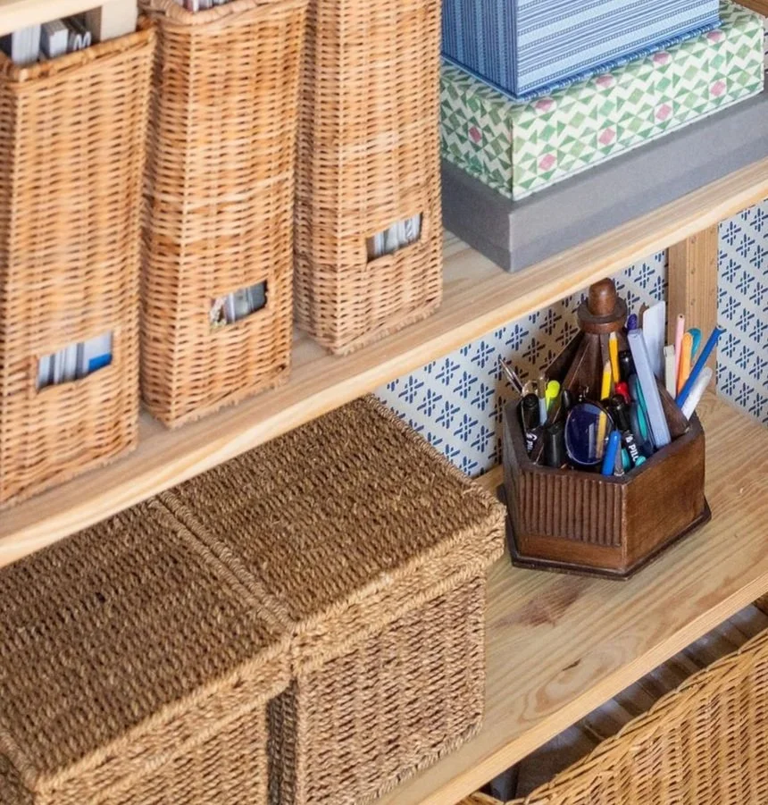 Wicker baskets and storage boxes on a wooden shelf, with a pen holder containing various writing instruments and a stack of decorative boxes or tissue boxes on top.