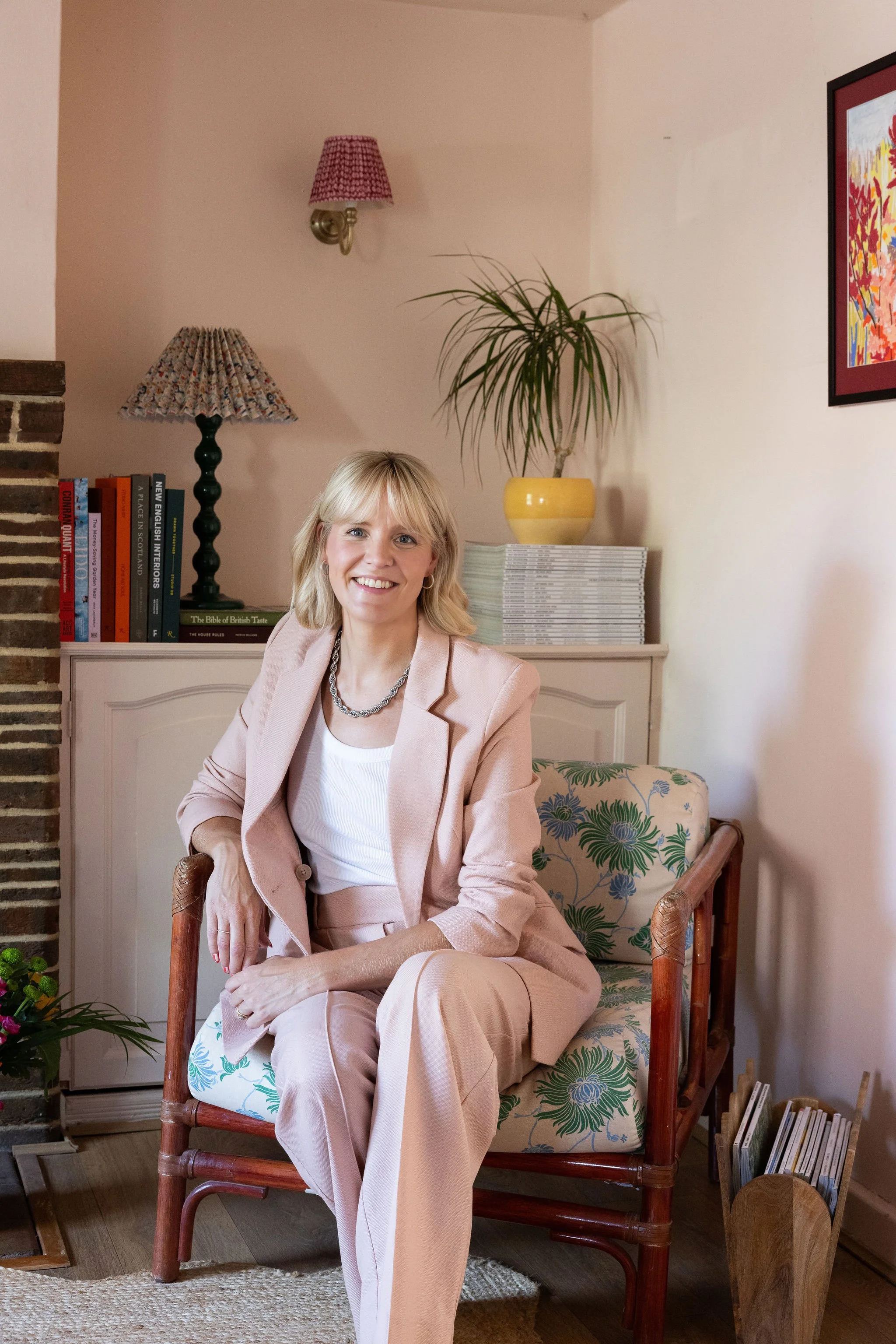 A woman with blond hair and a light pink suit sitting on a wooden chair with floral cushion in a living room. Behind her are bookshelves, a lamp with a patterned shade, and a potted plant. Part of a framed colorful painting is visible on the wall.