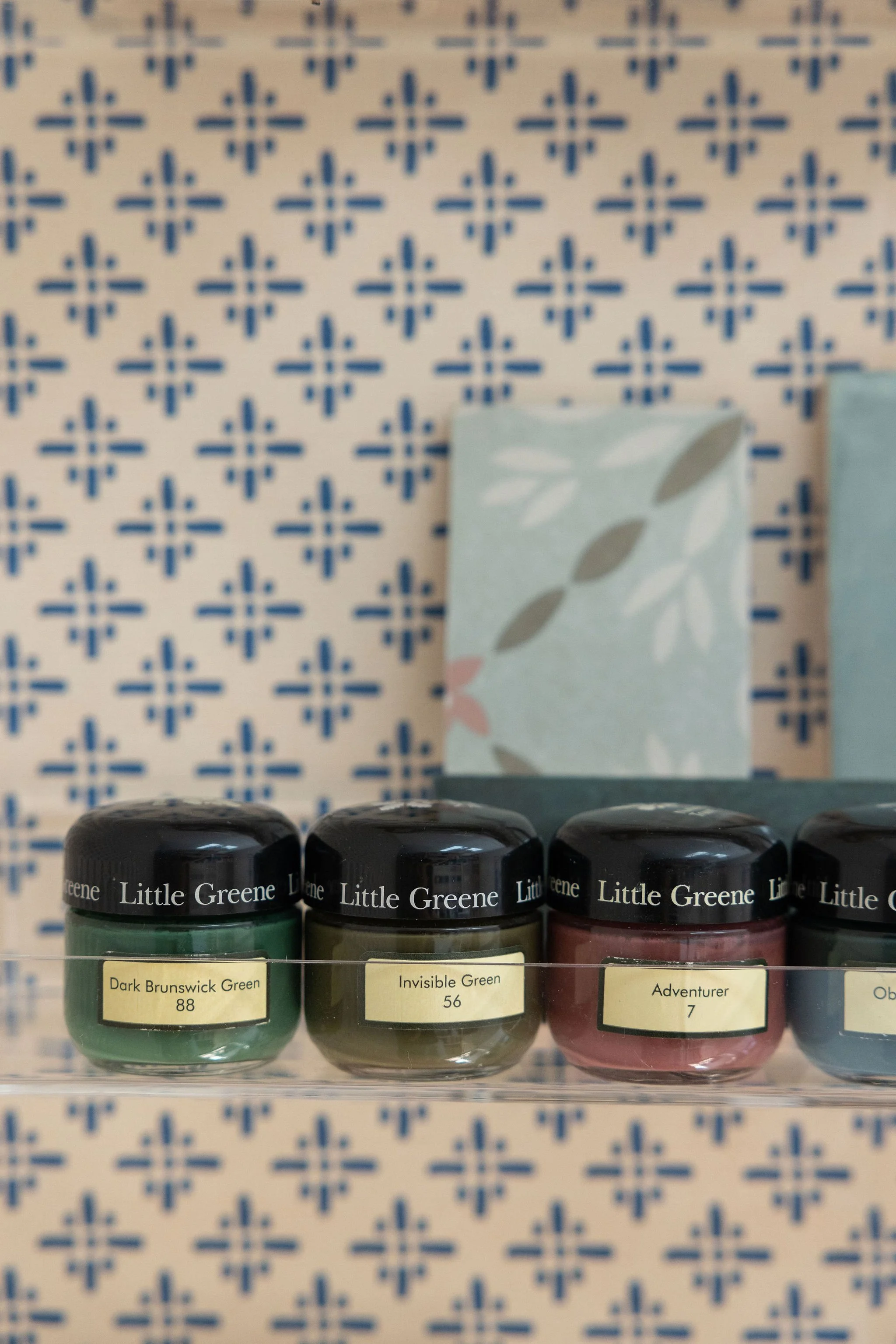 Close-up of small jars of waterproofing paint, labeled with colors 'Dark Brunswick Green,' 'Invisible Green,' and 'Adventurer,' with a patterned wall in the background.