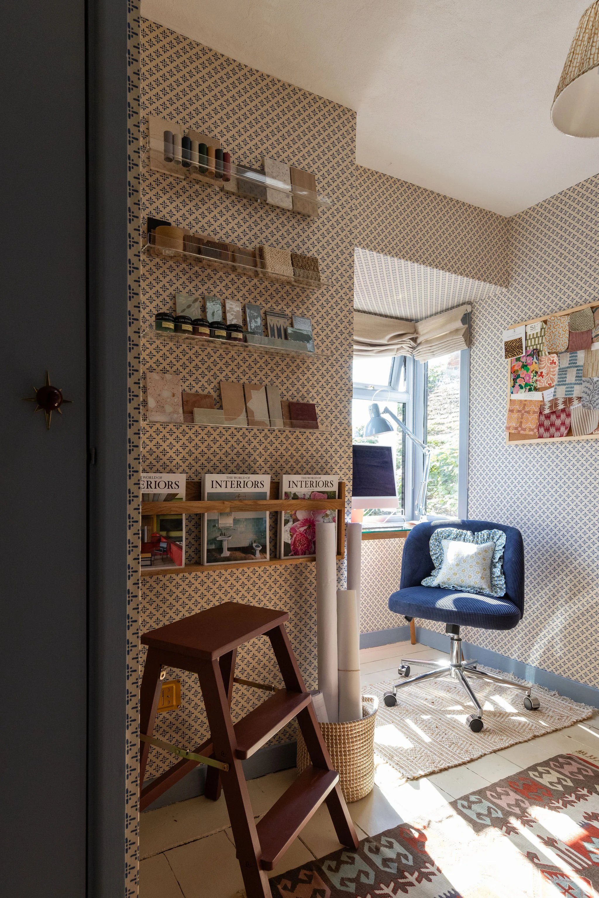 Home office with a blue swivel chair, a window with beige roman shades, patterned wallpaper, framed fabric swatches on the wall, a wooden step stool, a tall white candle, a small wooden ladder, a woven basket, a geometric-patterned rug, and assorted books and decor pieces on shelves.