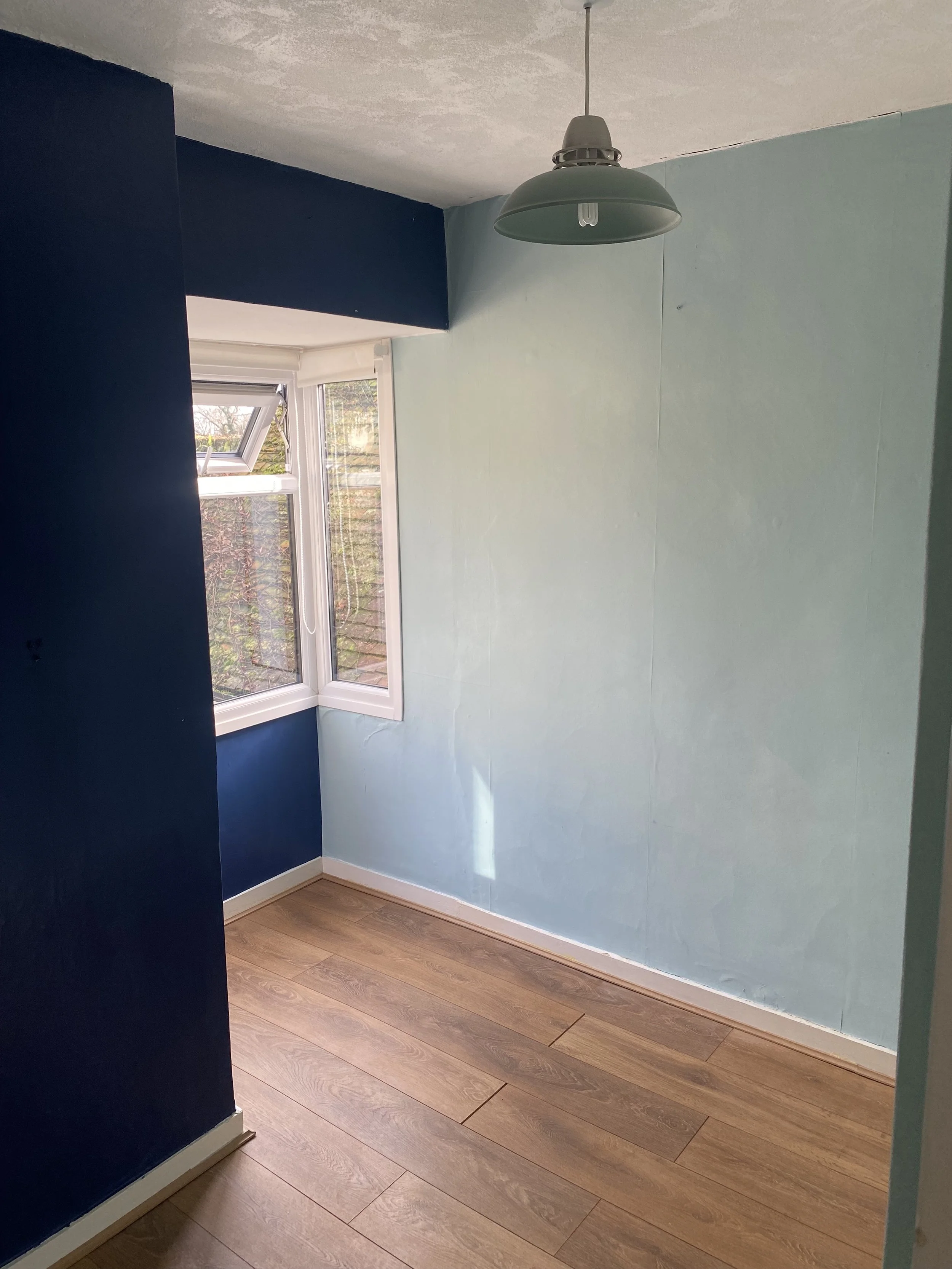 A room with one wall painted dark blue and the other light blue, a window with an open casement, a hanging light fixture, and wood flooring.
