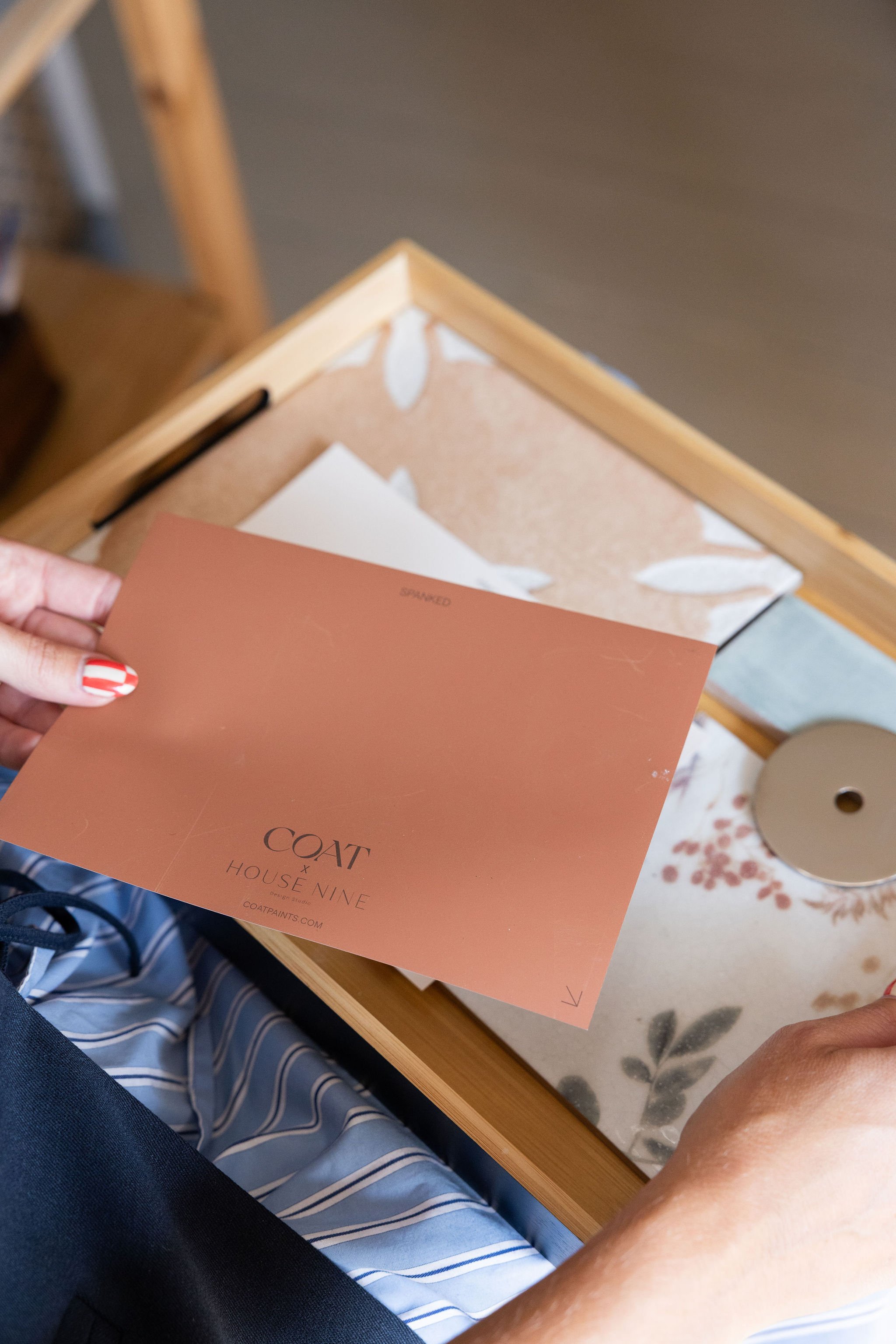 Close-up of a person's hand holding a peach-colored card with text "COAT HOUSE NINE" and website "COATPAINTS.COM." The background shows a wooden table, a sheet of paper, and surrounding fabric with blue and white stripes and a floral pattern.