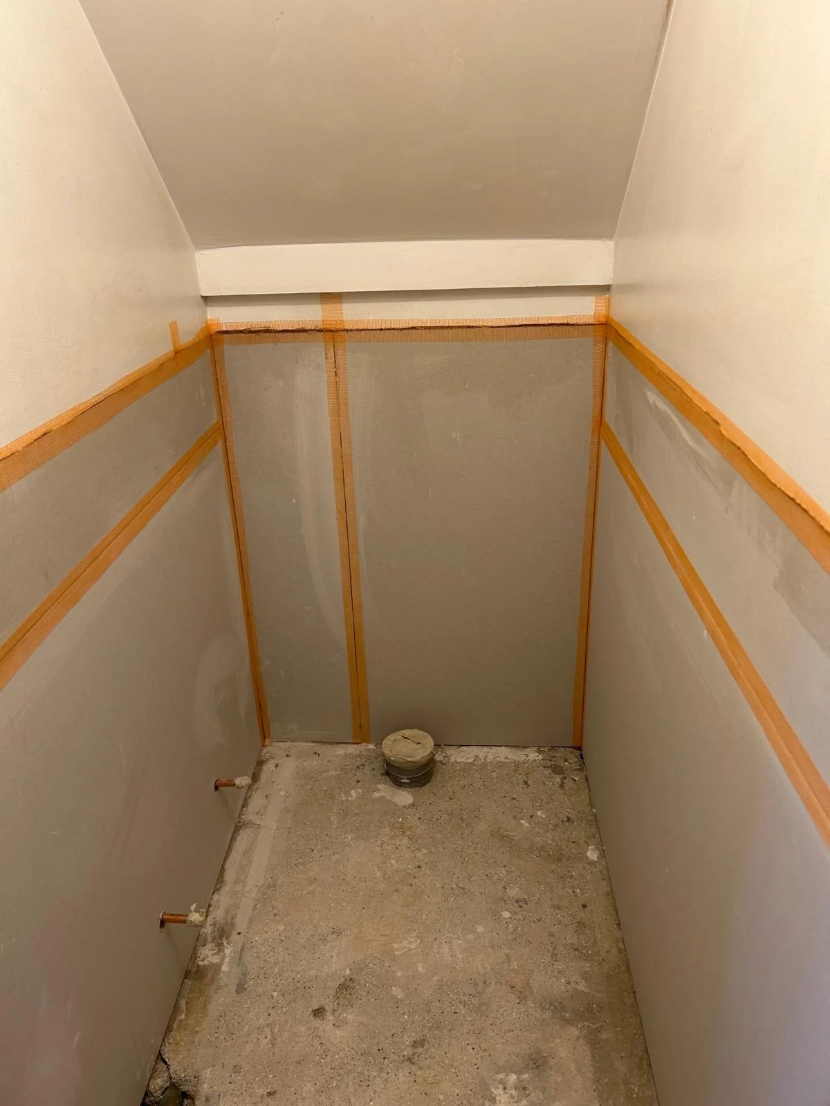 Construction of a small, unfinished bathroom or closet with partial drywall installed on the walls, orange tape outlining sections, and a round pipe or vent on the floor.