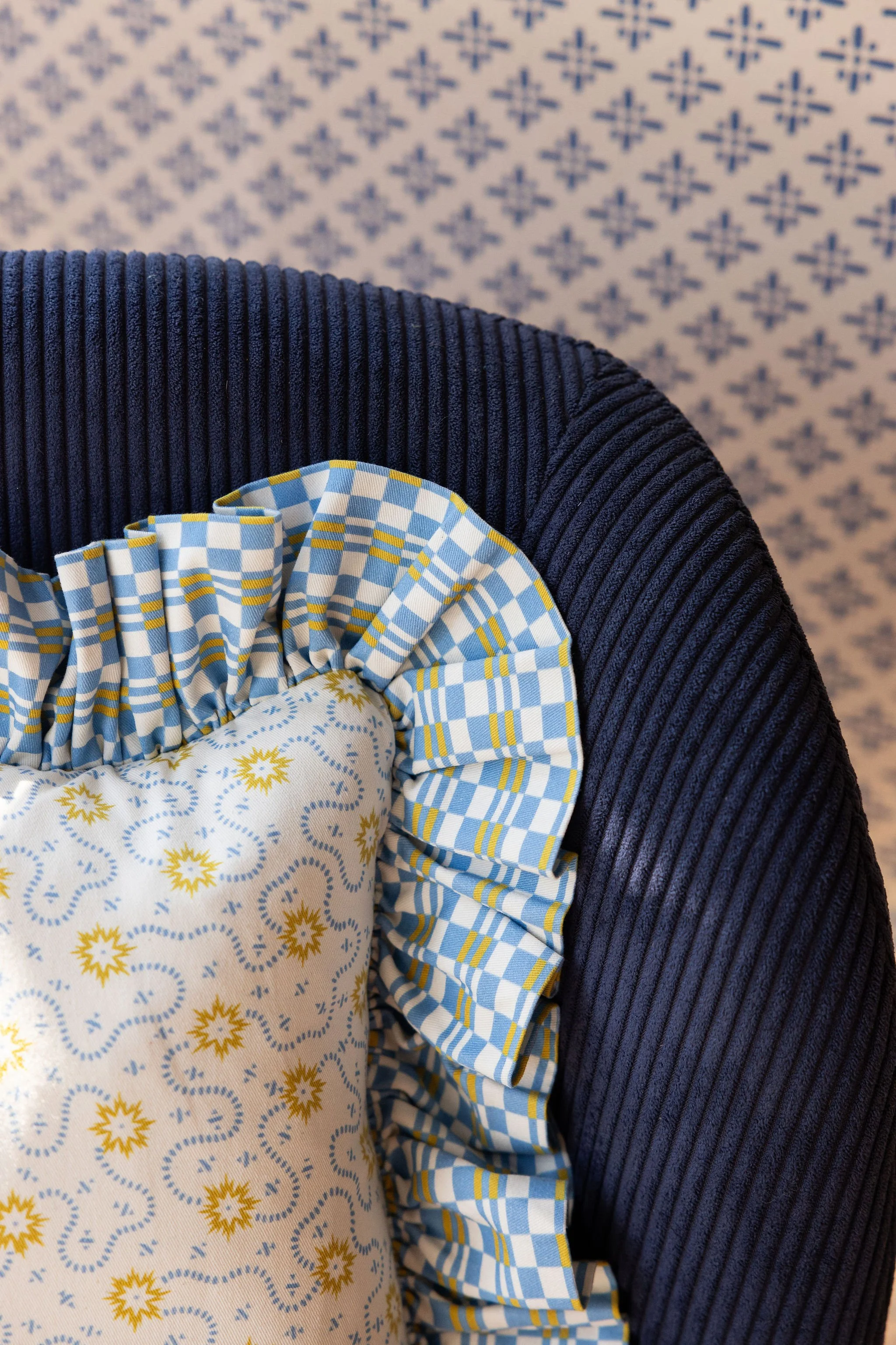 Close-up of a chair with a blue and white checked ruffle and a patterned cushion with yellow sun and star motifs, placed on a dark blue, textured armrest against a background with a blue geometric wall pattern.