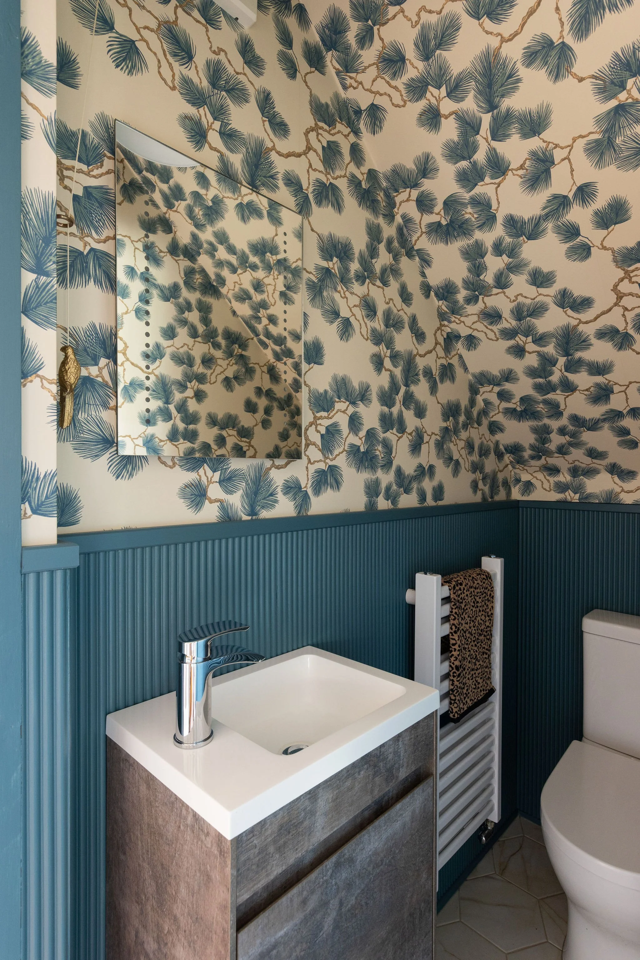Small modern bathroom with beige wallpaper featuring blue palm leaves and brown branches, blue beadboard wainscoting, a white vanity with a textured gray cabinet, a rectangular sink, a sleek chrome faucet, a mirror, a towel radiator with a leopard print towel, and part of a toilet visible.