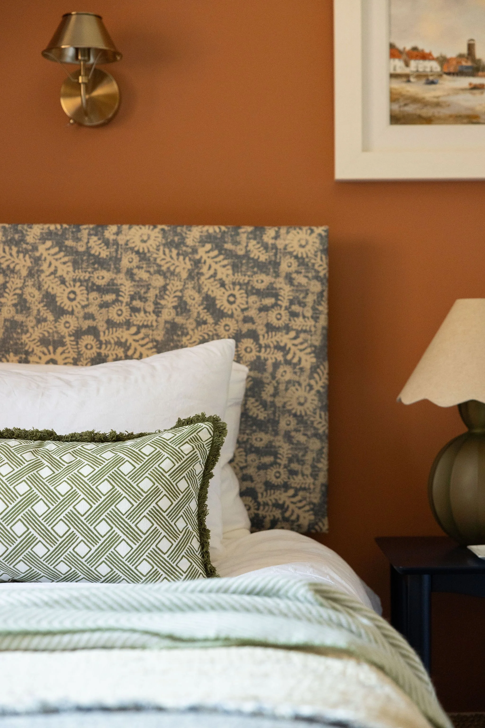 A neatly made bed with a patterned green and white pillow, white linens, and a bed runner with green and white geometric pattern. The headboard has a blue and white floral pattern. To the right, a side table holds a lamp, and a framed picture of a coastal town hangs on the earth-colored wall. A brass wall-mounted light fixture is above the bed.