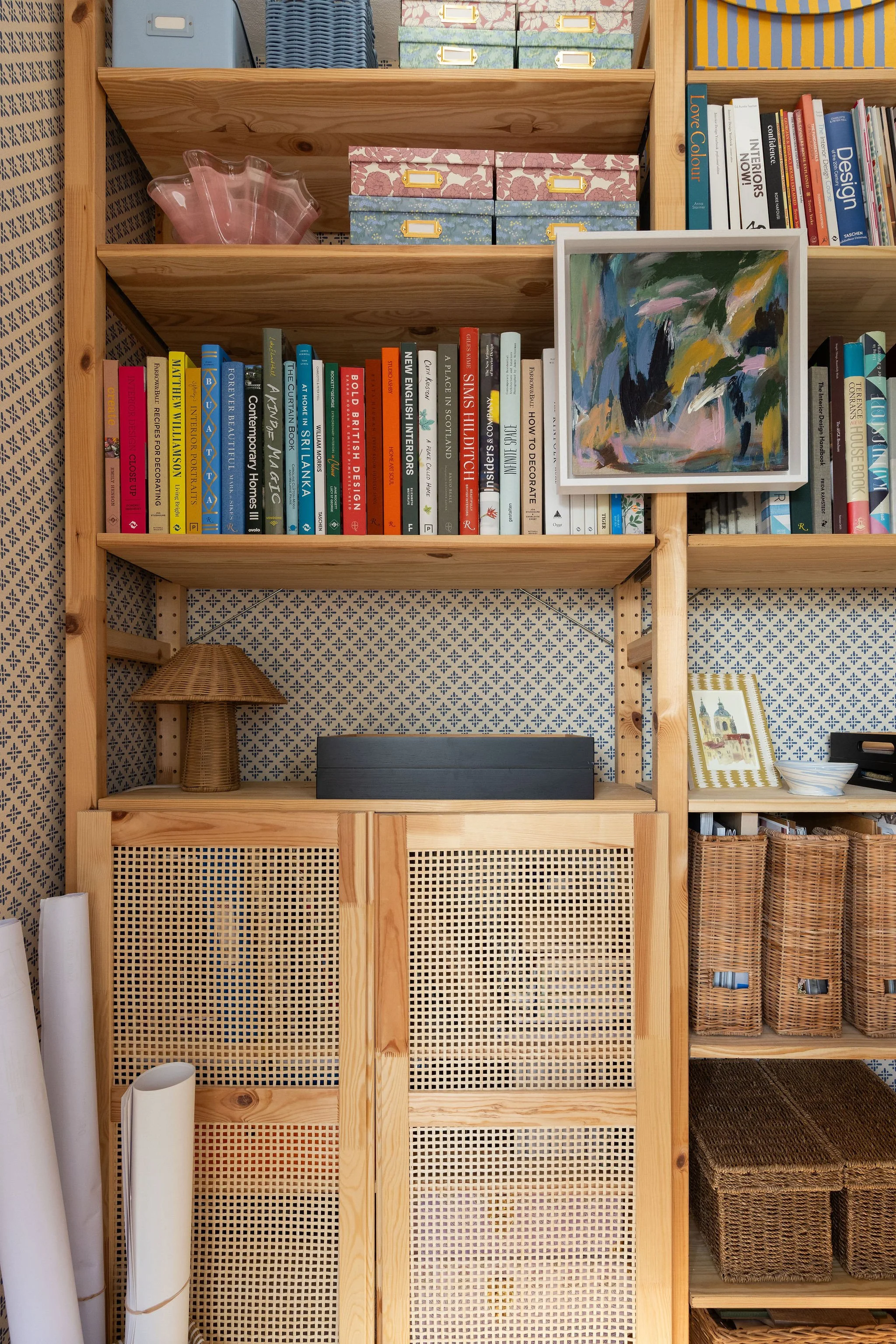 A wooden shelving unit with decorative storage boxes, books, artwork, a pink glass dish, a small wicker table lamp, and baskets stored on the shelves.