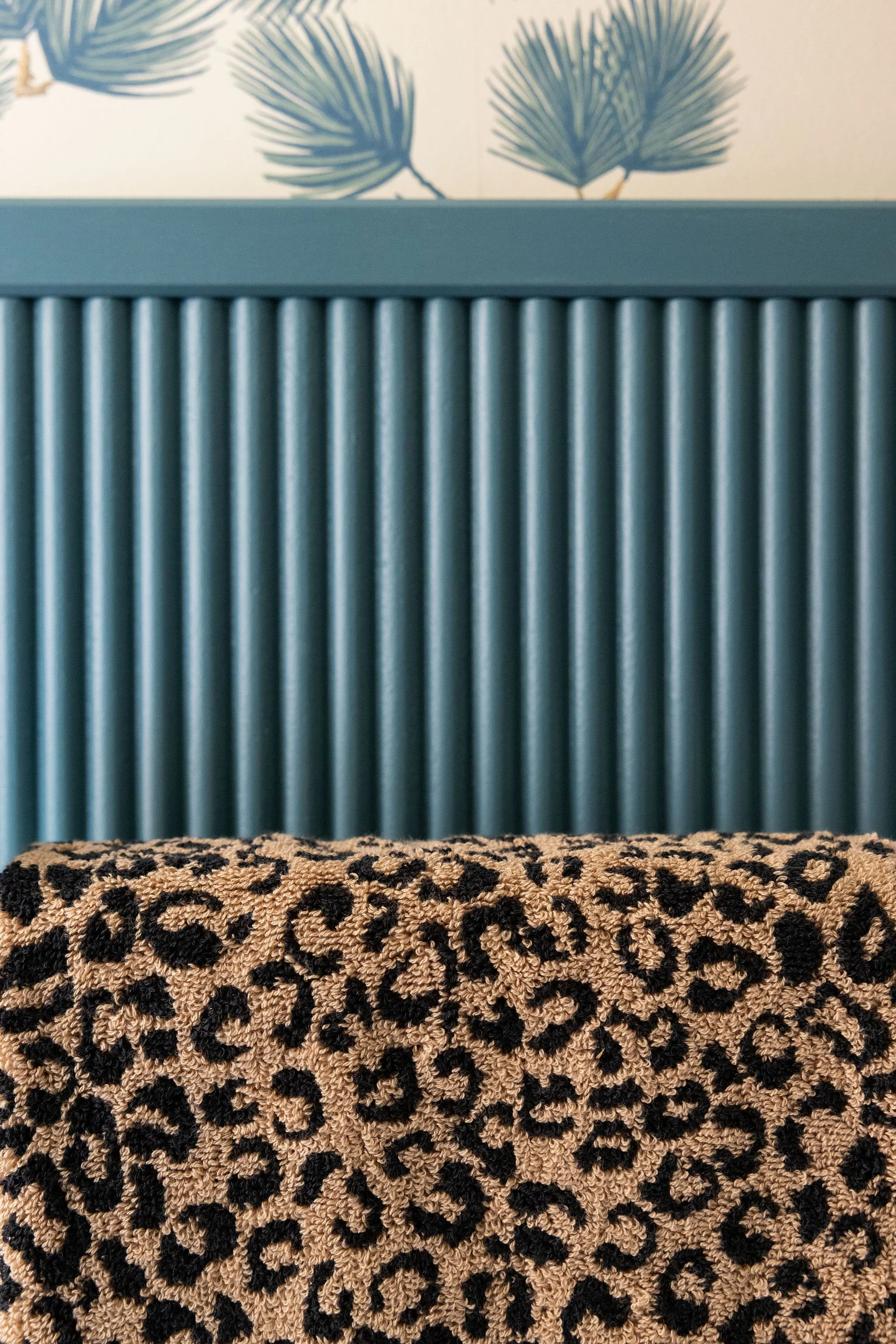 Partial view of a teal-colored radiator, wallpaper with palm leaves, leopard-print textured fabric on a surface in the foreground.