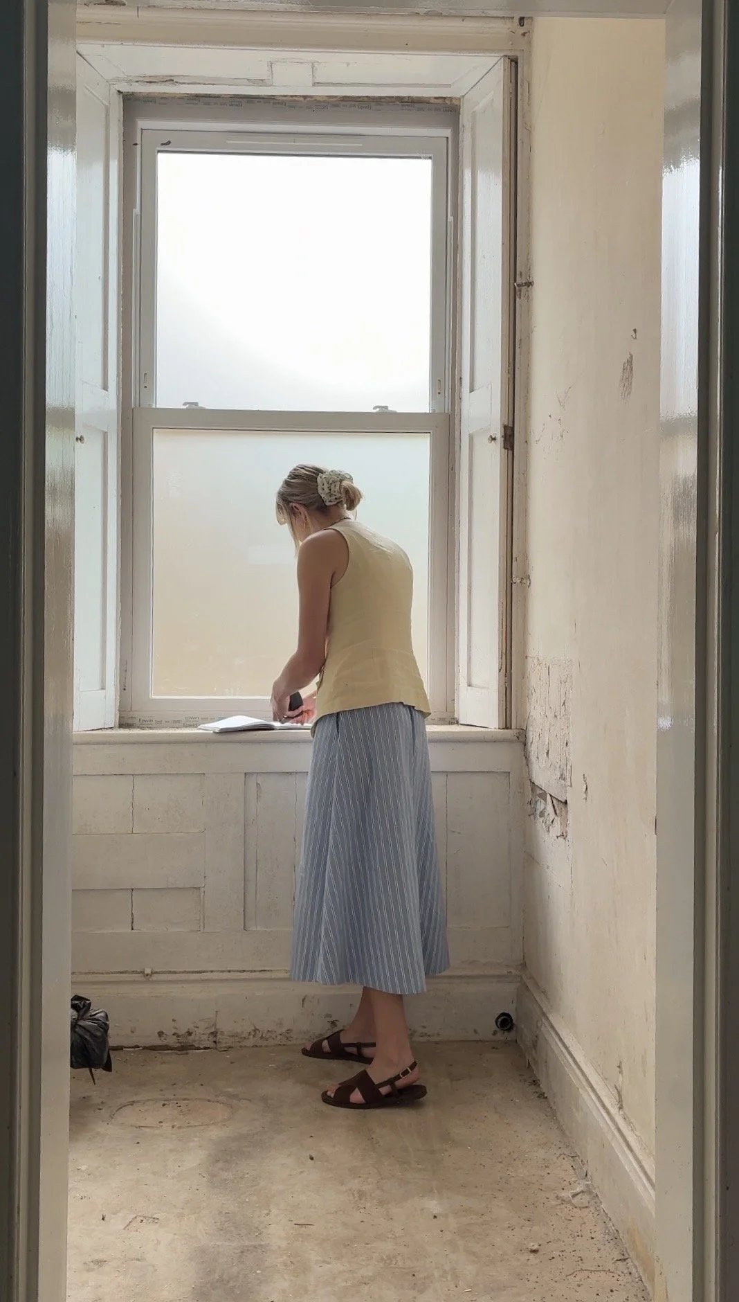 A woman in a yellow sleeveless top and blue striped skirt standing in a neglected room by a window, looking at something on the windowsill.