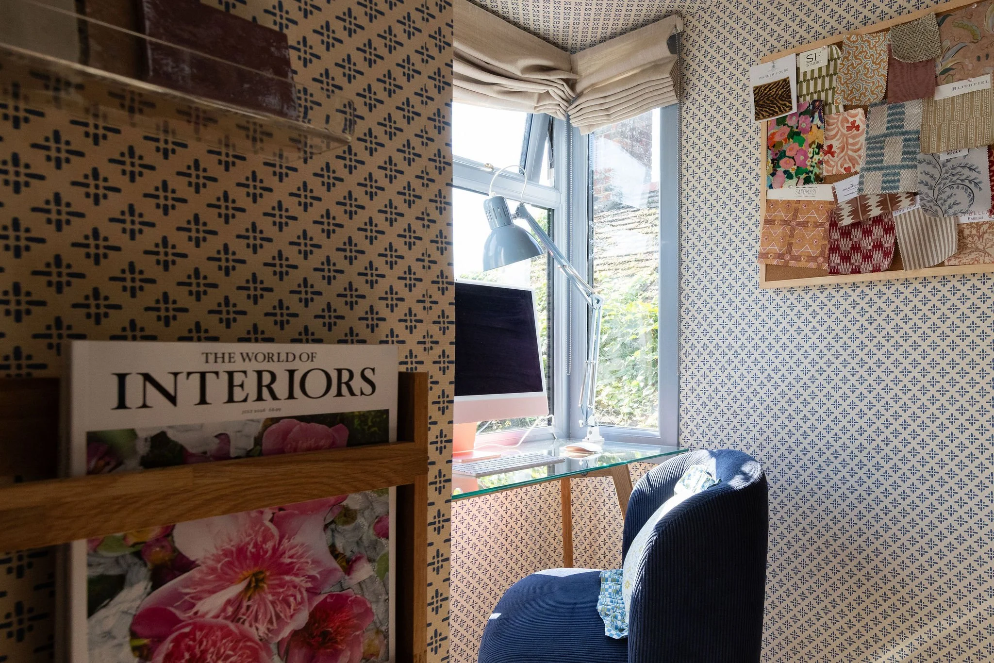 An interior room corner with patterned wallpaper, a window with draped curtains, a desk lamp, a computer, decorative fabric samples on a wall, and a bookshelf with a magazine titled 'The World of Interiors'.