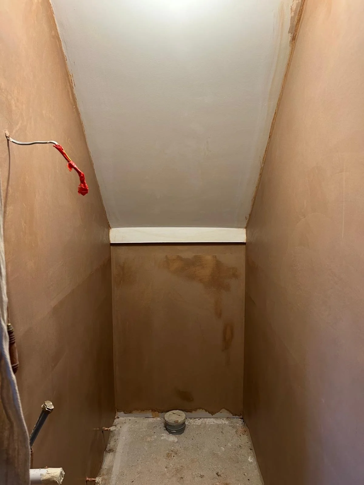 An unfinished small bathroom with drywall and plumbing, with a pipe at the floor and wiring on the wall, awaiting further construction.