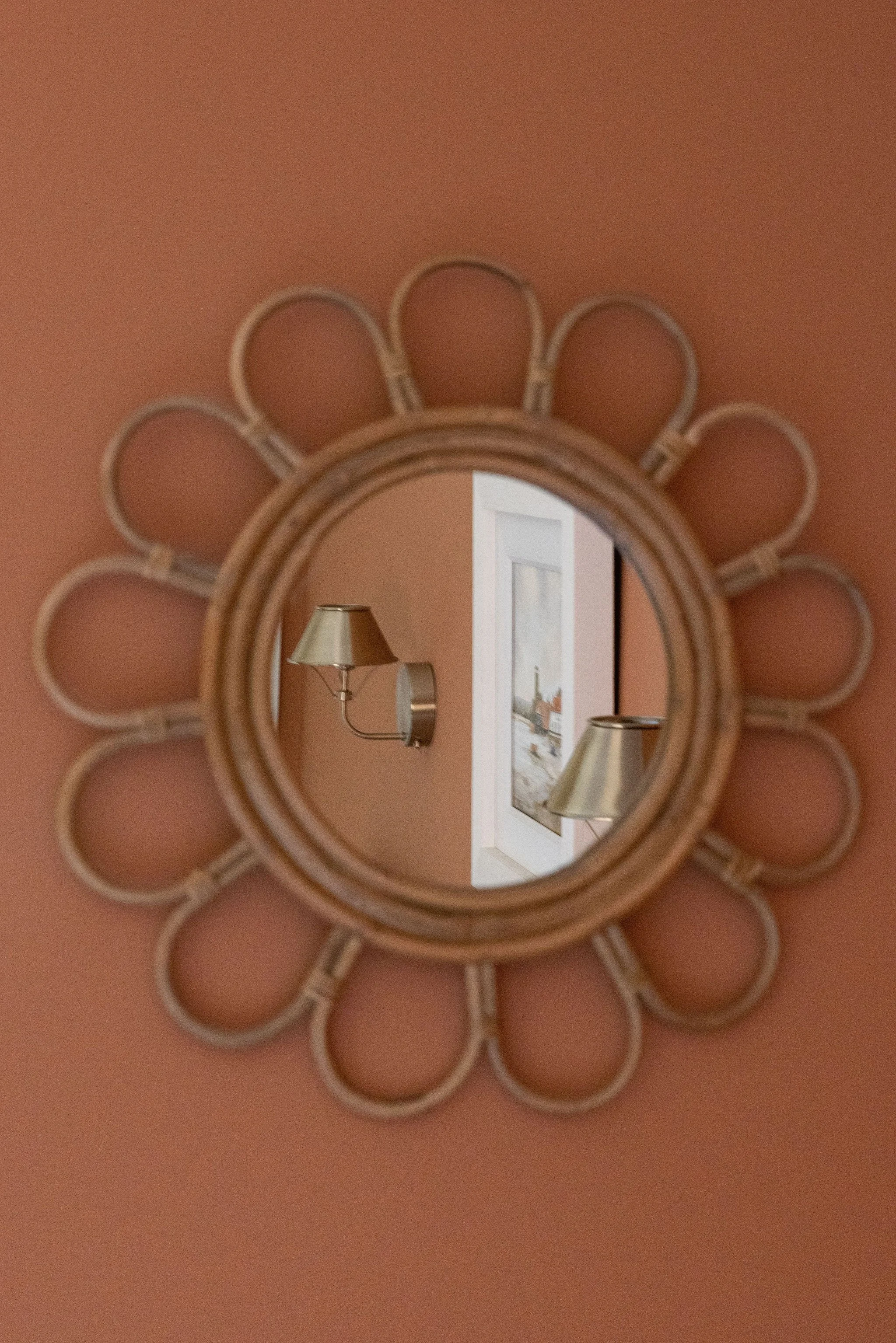 Round mirror with a decorative frame hanging on a peach-colored wall, reflecting a wall sconce lamp and a framed picture.
