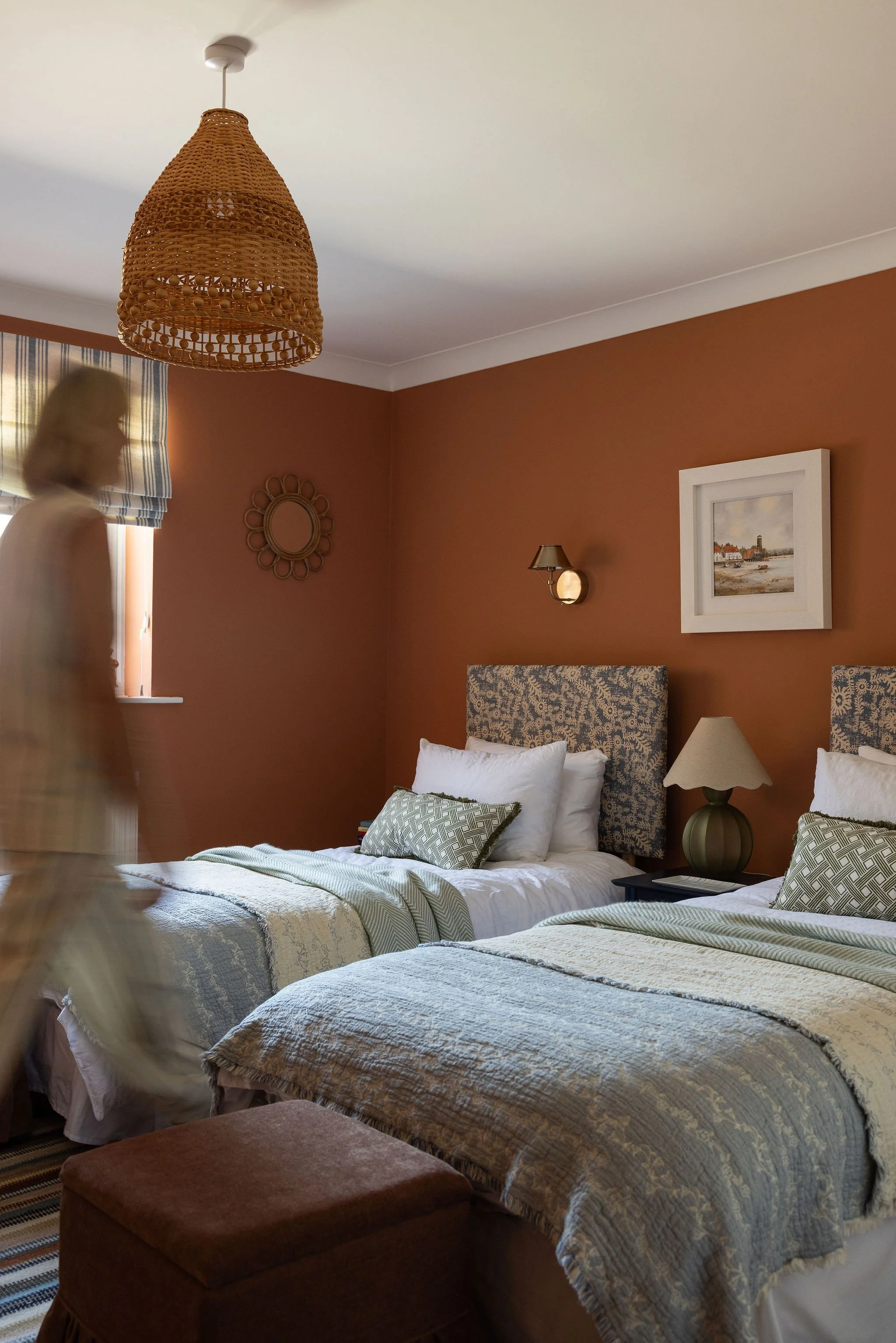 Two beds with patterned bedspreads and pillows in a room with warm-colored walls. A bedside table with a lamp and framed artwork on the wall are visible. A blurred person is walking past the room.