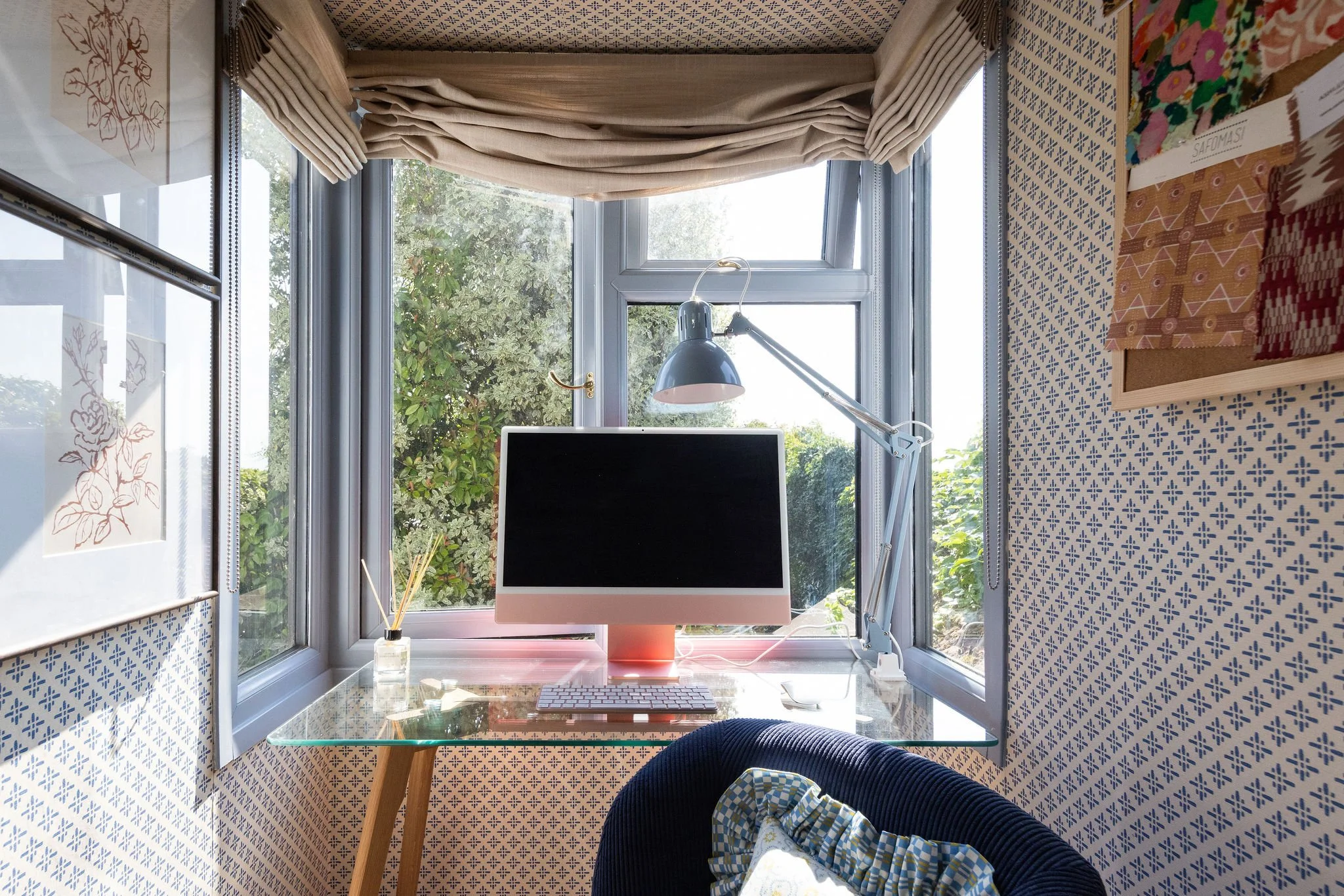 A home office corner with a desk, computer monitor, lamp, and decor. The window has beige curtains and shows outdoor greenery. The wall features patterned wallpaper and framed artwork.