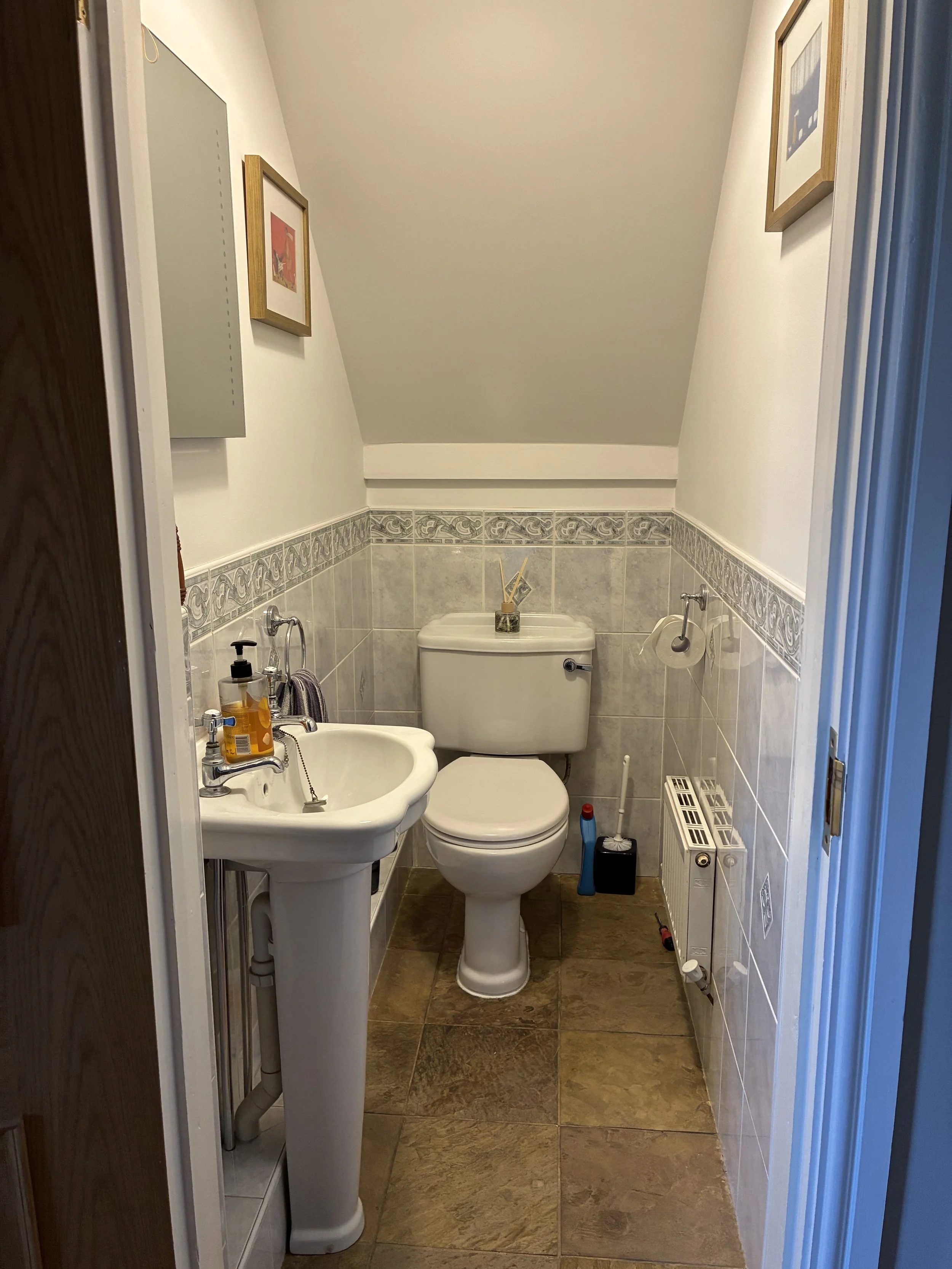 A small bathroom with a toilet, a sink, and cleaning supplies, with framed artwork hanging on the walls and beige tiled flooring.