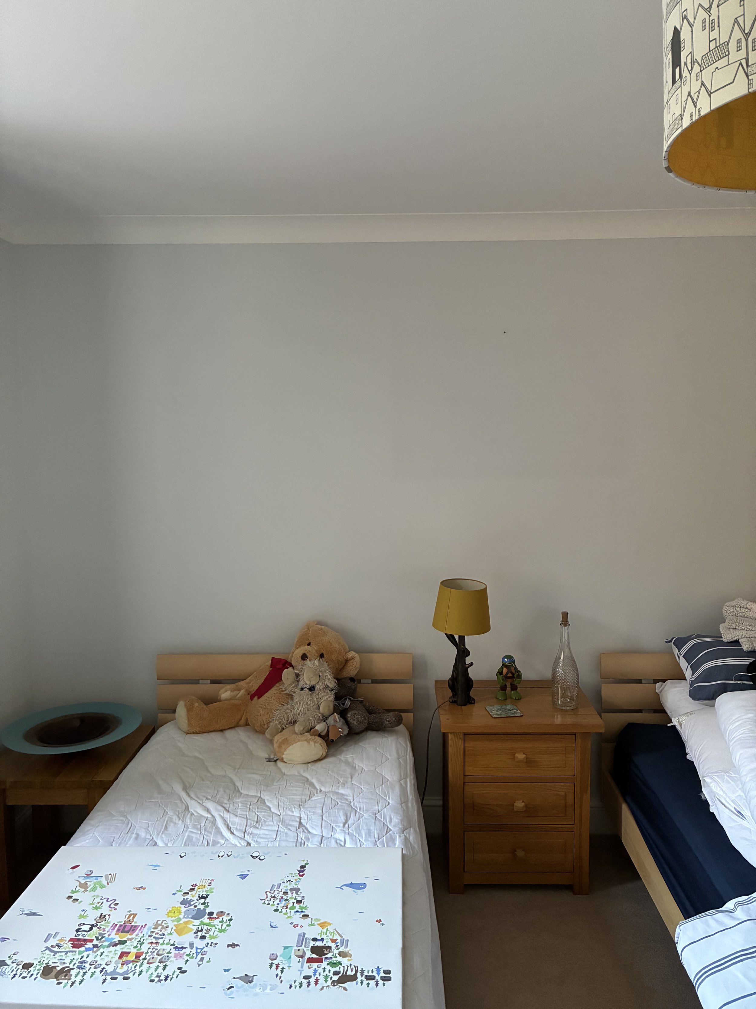 A bedroom with a wooden bed, stuffed animals, a nightstand with a lamp and figurines, a second bed with striped bedding, and a white board game with colorful illustrations on top