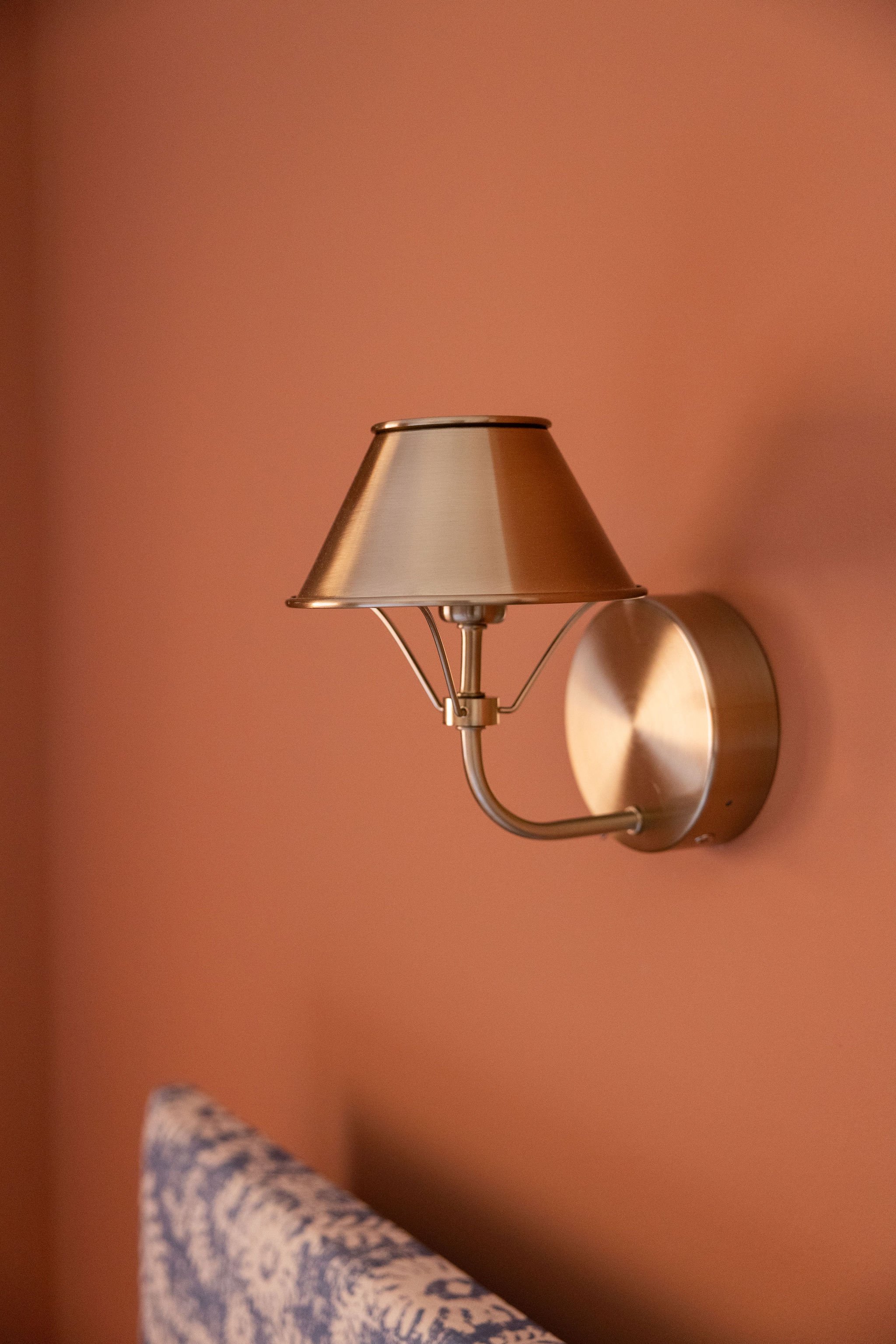 A metallic wall-mounted reading lamp with a conical shade and an adjustable arm, attached to a pink wall. Part of a person's arm wearing a patterned blue and white garment is visible in the lower left corner.