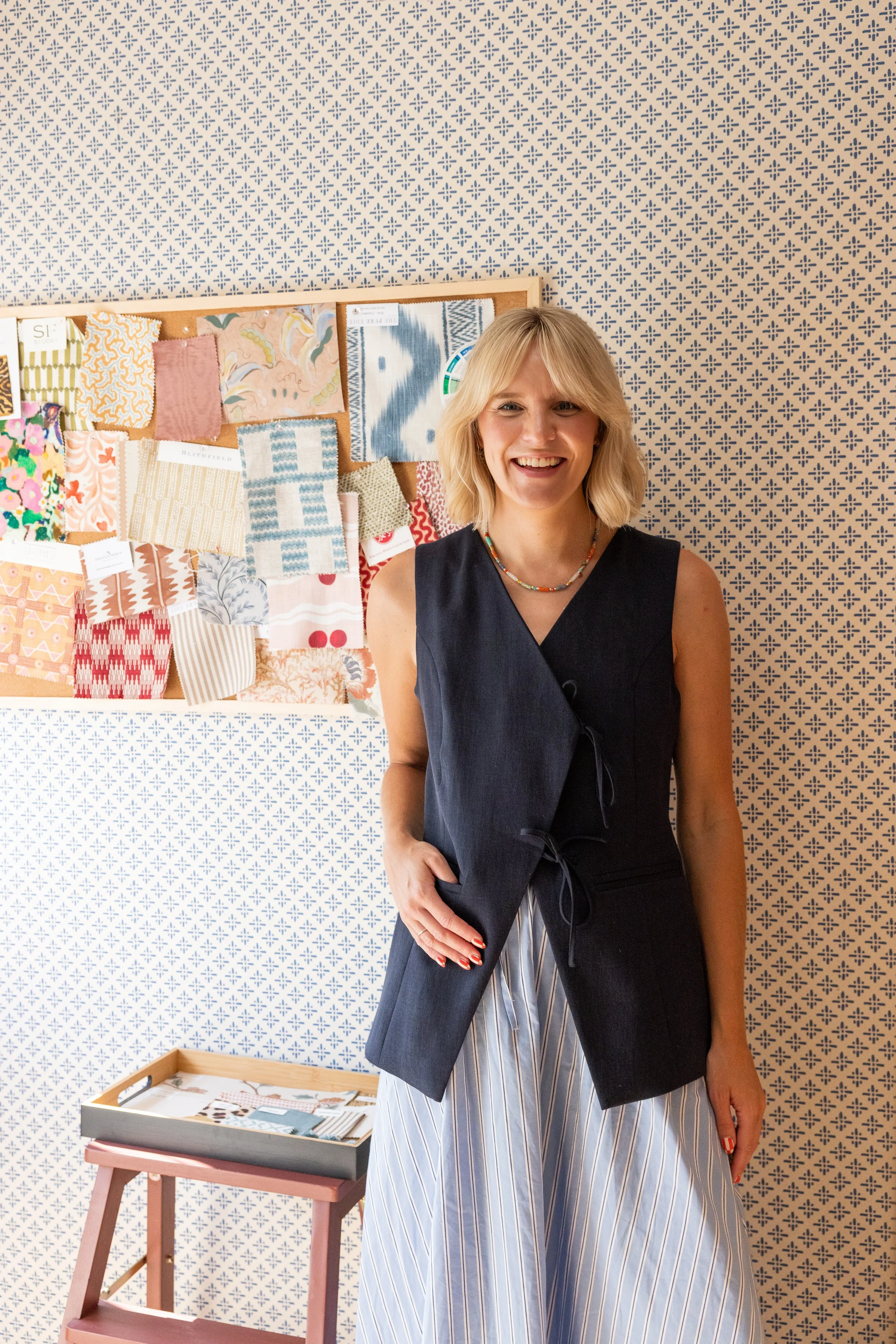A smiling woman with blonde hair, wearing a blue sleeveless top, a colourful beaded necklace, and striped wide-leg skirt, standing in a room with a patterned wallpaper and a fabric swatch display board background.