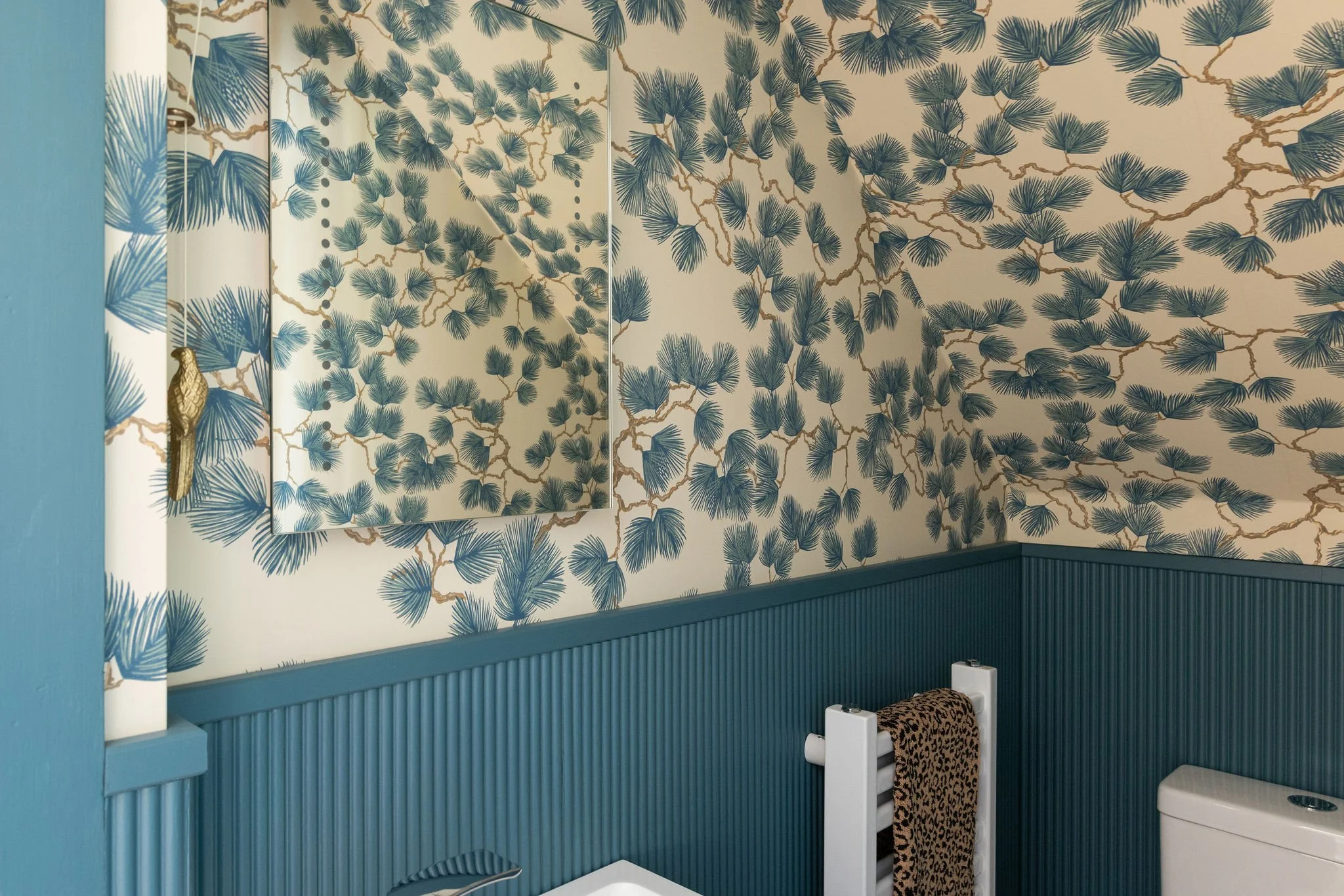 Interior of a room with a blue color scheme, featuring a wall with Tommy Bahama tropical palm tree wallpaper, a mirror, a white radiator with a leopard print towel, and a white toilet.