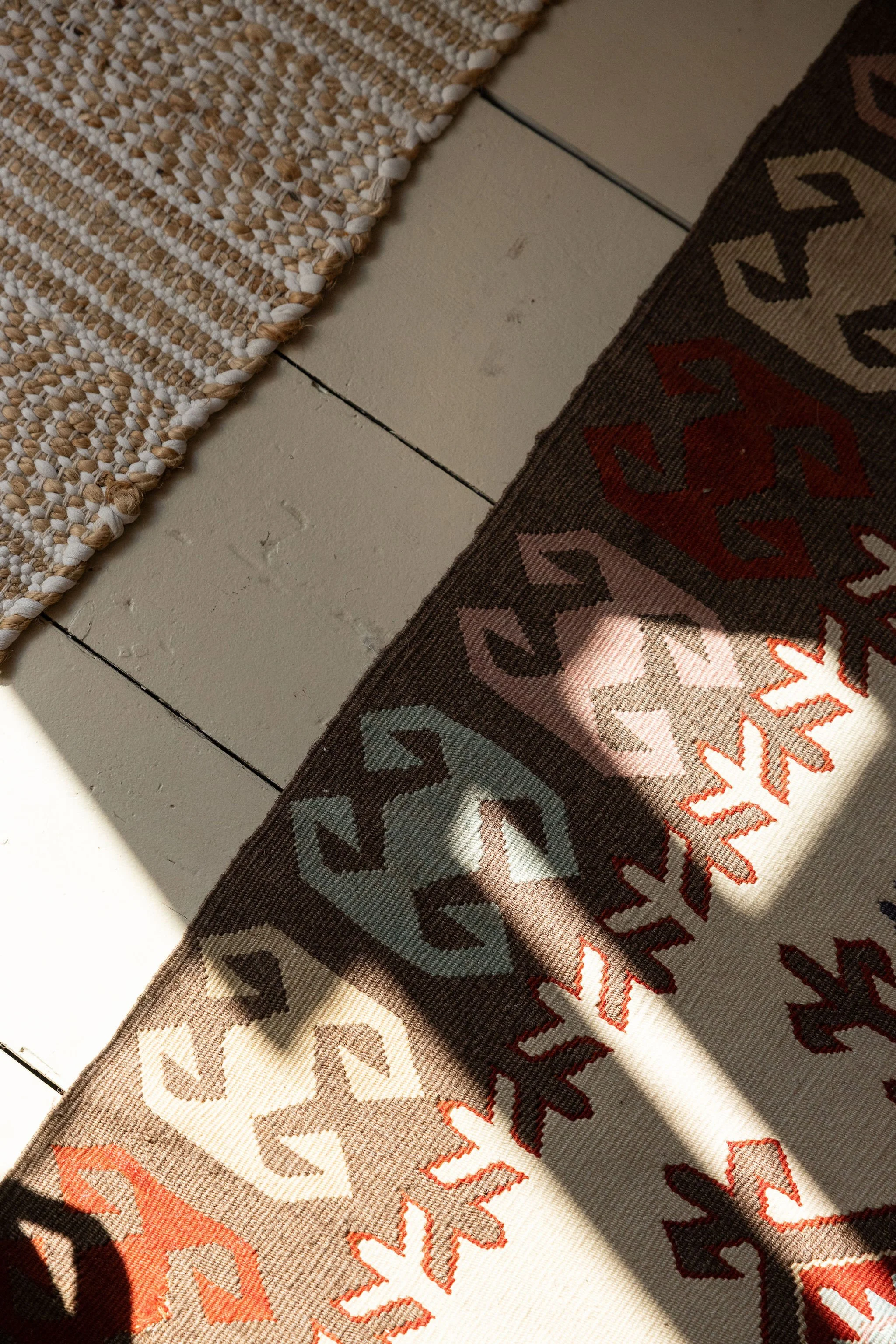 Close-up of a textured woven rug with a pattern of geometric shapes in beige, brown, and reddish-orange colors, laid on a white painted wooden floor.