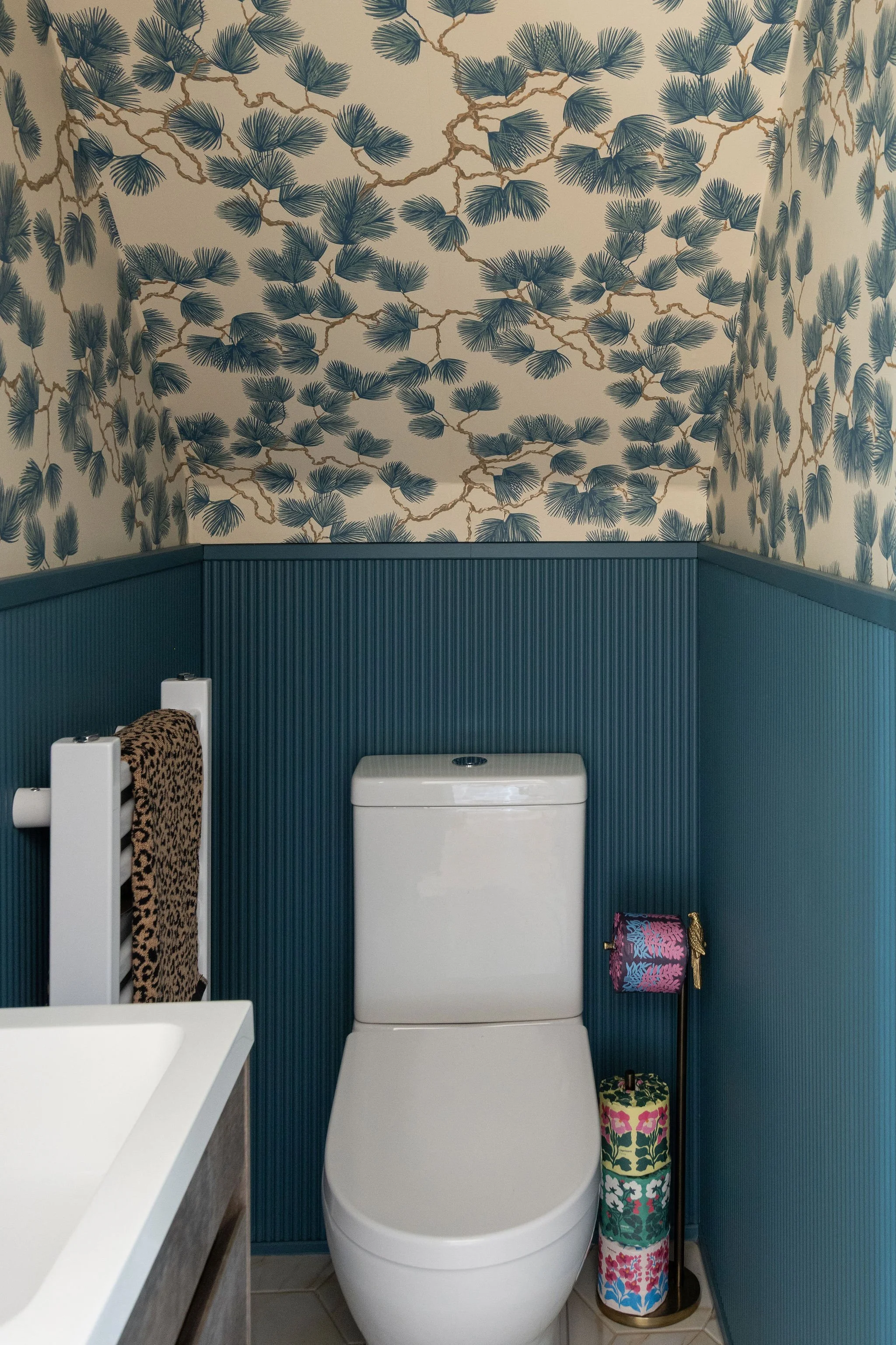 A bathroom with a modern white toilet, a dark teal wainscot, patterned wallpaper with blue pine branches, a toilet paper stand with floral design, and a leopard print towel hanging on a white towel rack.