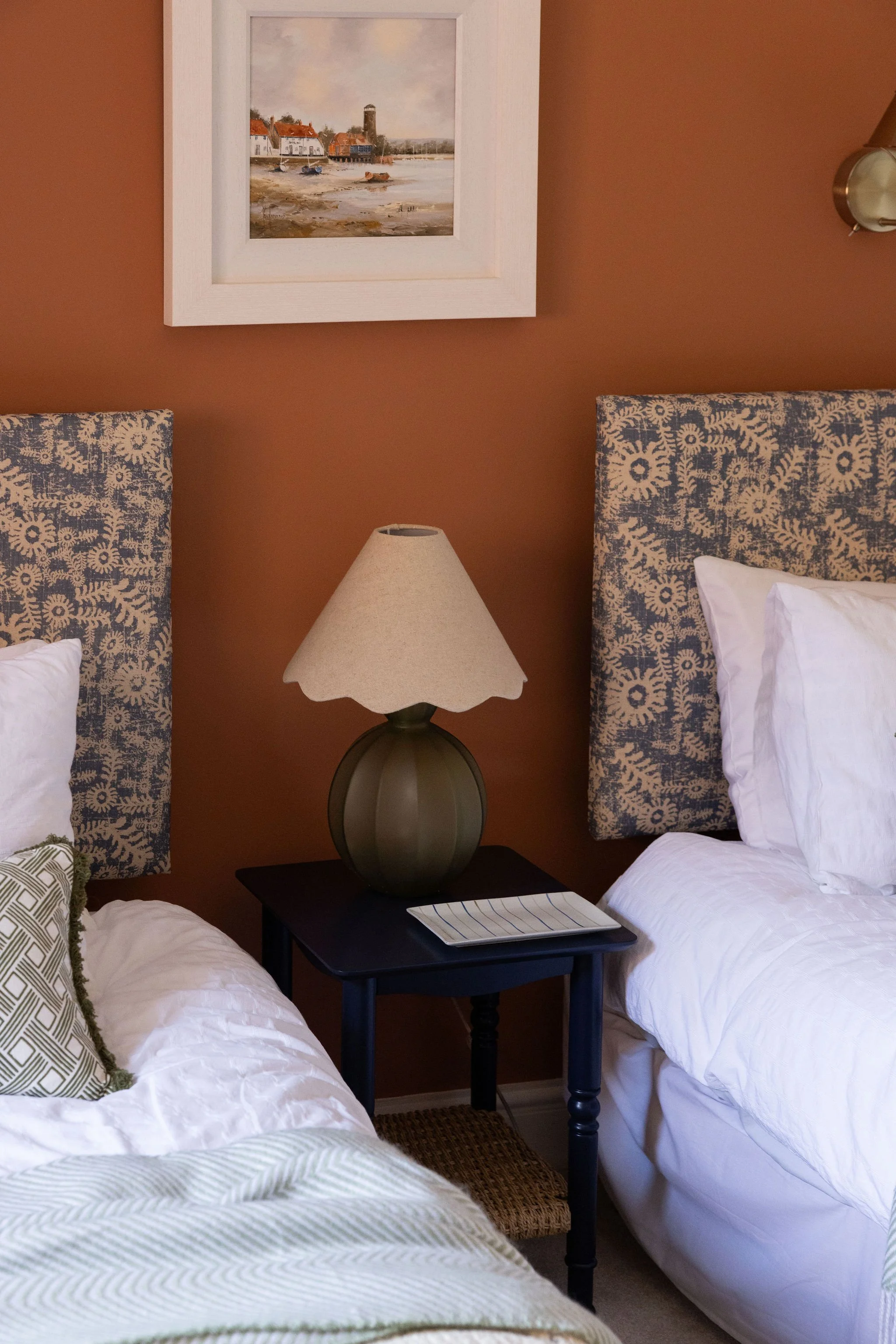 A bedroom with two single beds, each with white bedding and patterned pillows, flanking a black nightstand. The nightstand has a beige lampshade on a dark base and a striped tablet or book. Above, a framed painting of a waterfront scene with houses, 