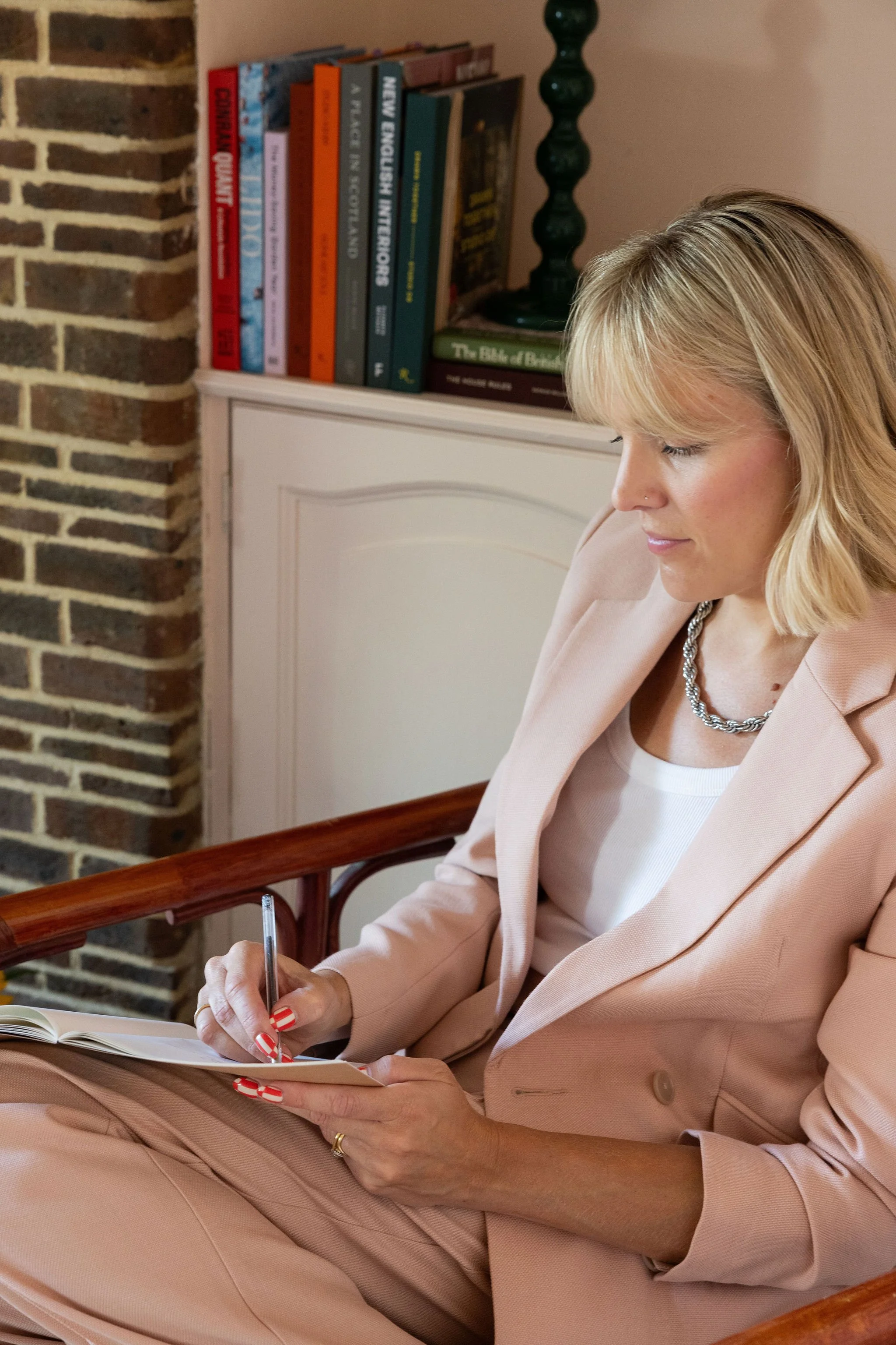 A woman with blonde hair and a nose piercing wearing a light pink blazer and pants, sitting and writing in a notebook with a small pen. She has red and white striped nails, and there is a white cabinet and a bookshelf in the background.