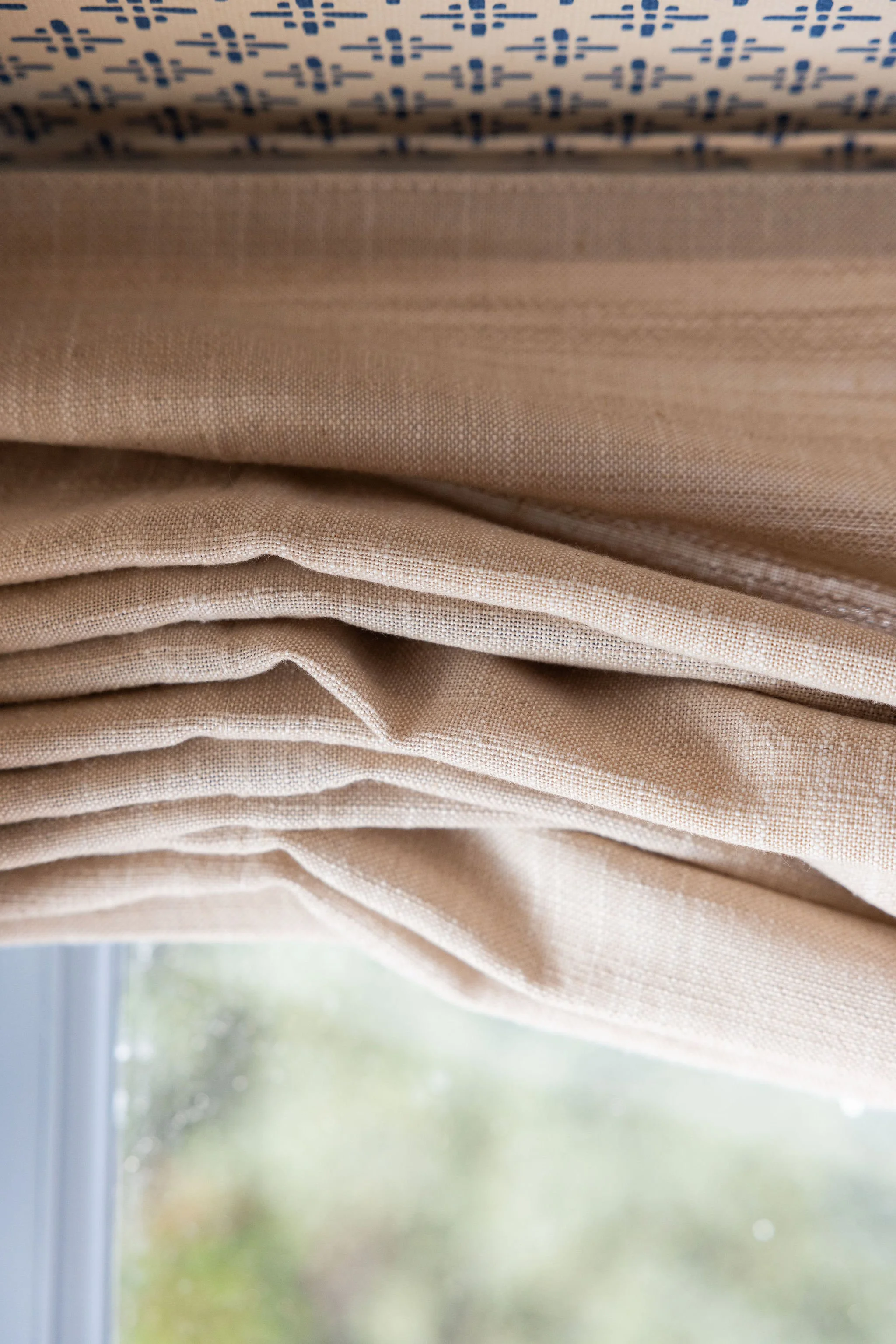 Close-up of beige linen curtains hanging in front of a window, with blurred outside view of trees and sky.