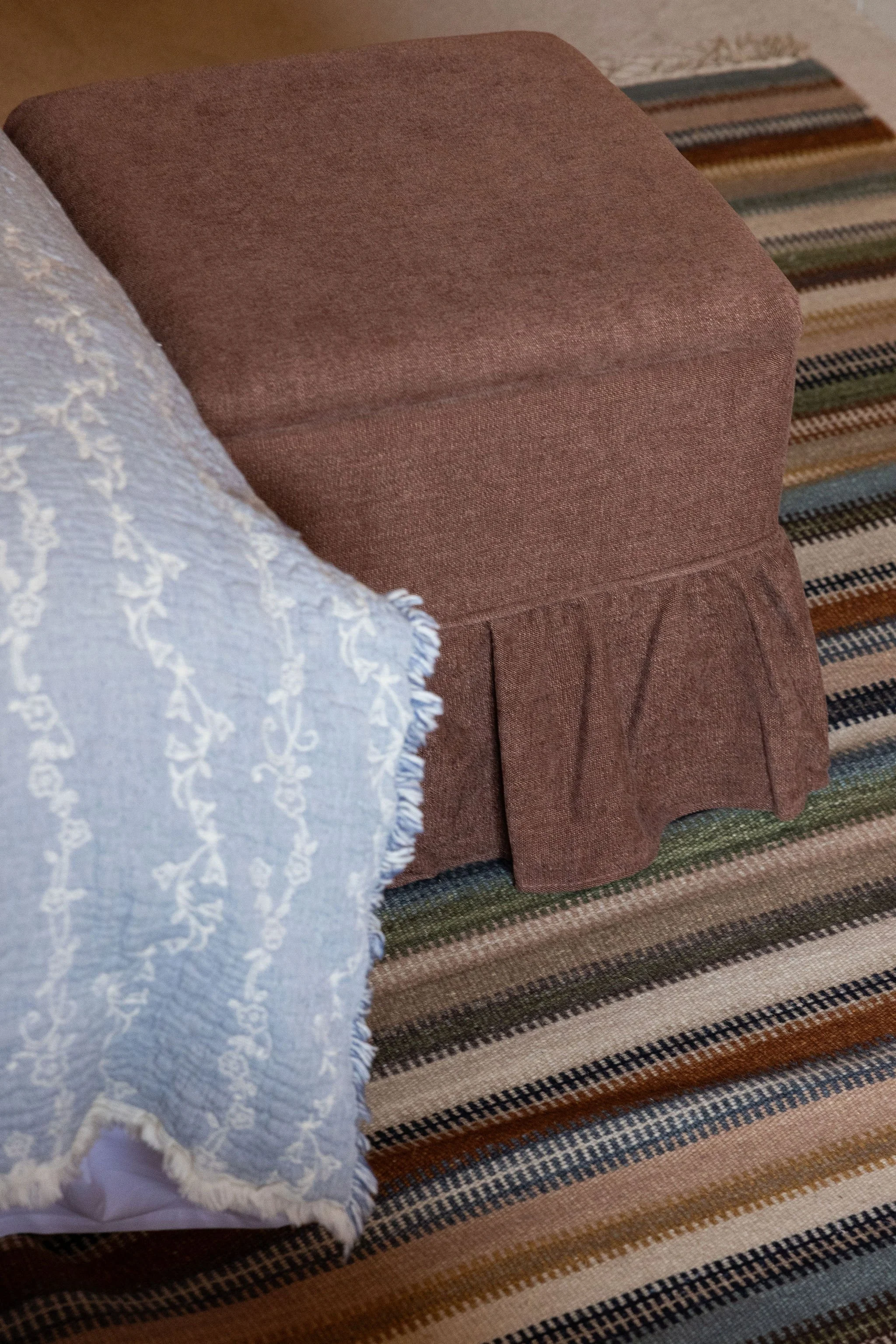 Close-up of a brown upholstered pouffe with a blue and cream embroidered cloth draped over it, on a striped rug with shades of brown, blue, and orange.